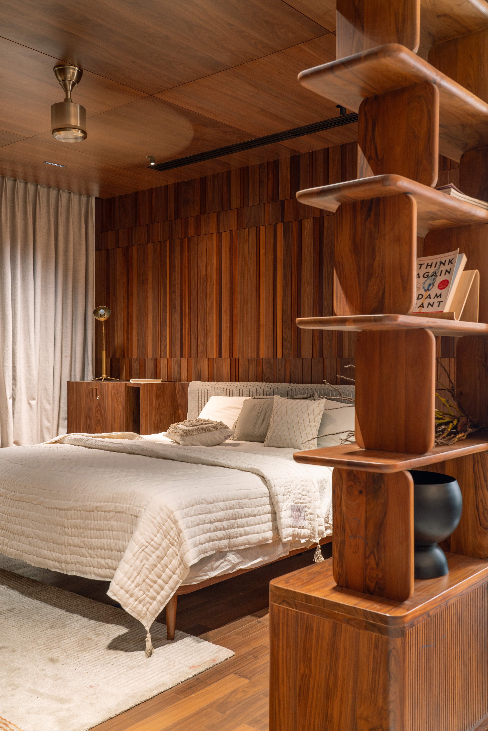 Bedroom clad in vertical teak slats with a sculpted timber shelving column beside a bed in warm lamplight