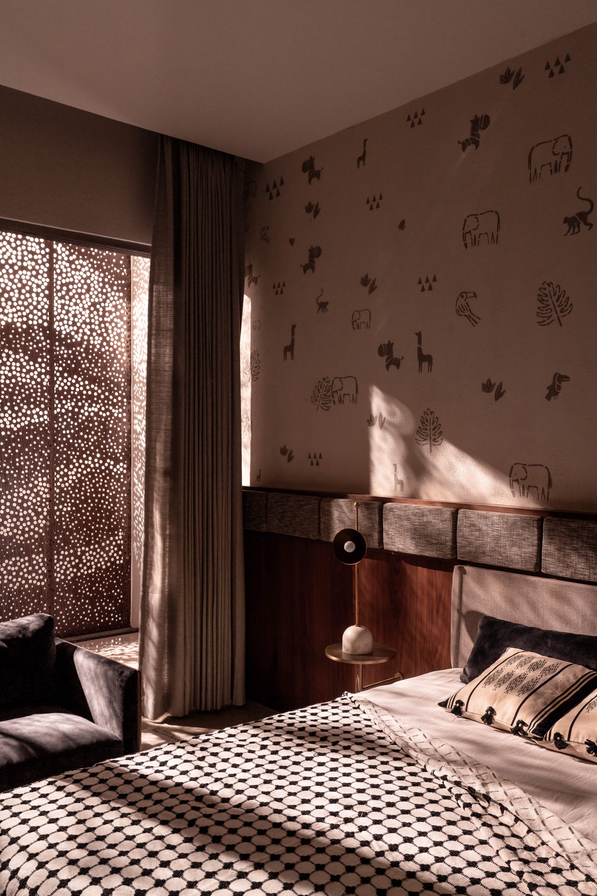 Bedroom wall printed with animal and leaf motifs beside a perforated screen, with afternoon sun falling across a checked bedspread