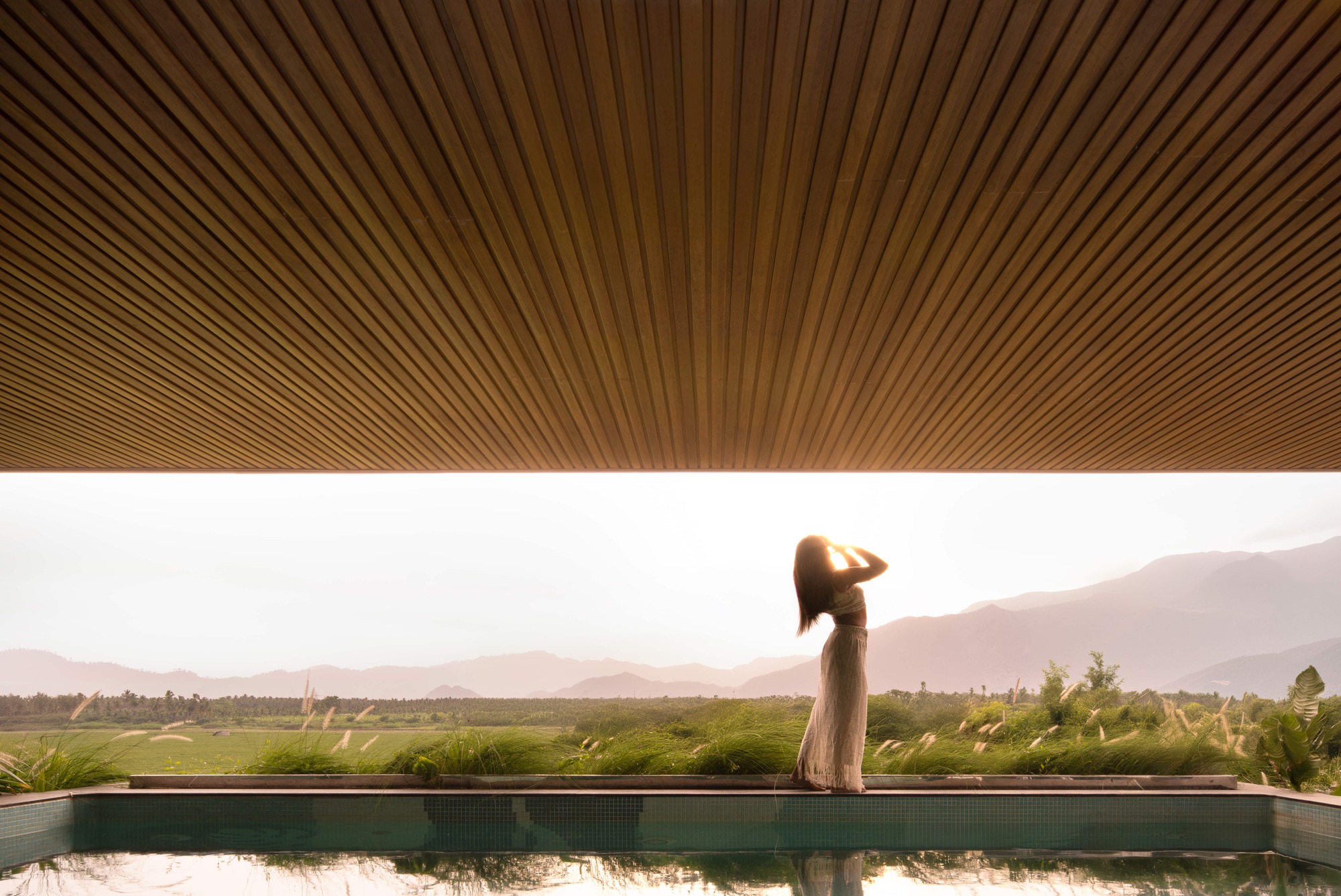 Slatted timber ceiling over an infinity pool edge, with a woman silhouetted against hazy mountains and paddy fields at sunrise