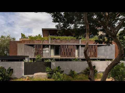 Street facade of weathered steel screens and grey concrete walls with a planted roof edge, framed by tree trunks under a cloudy sky