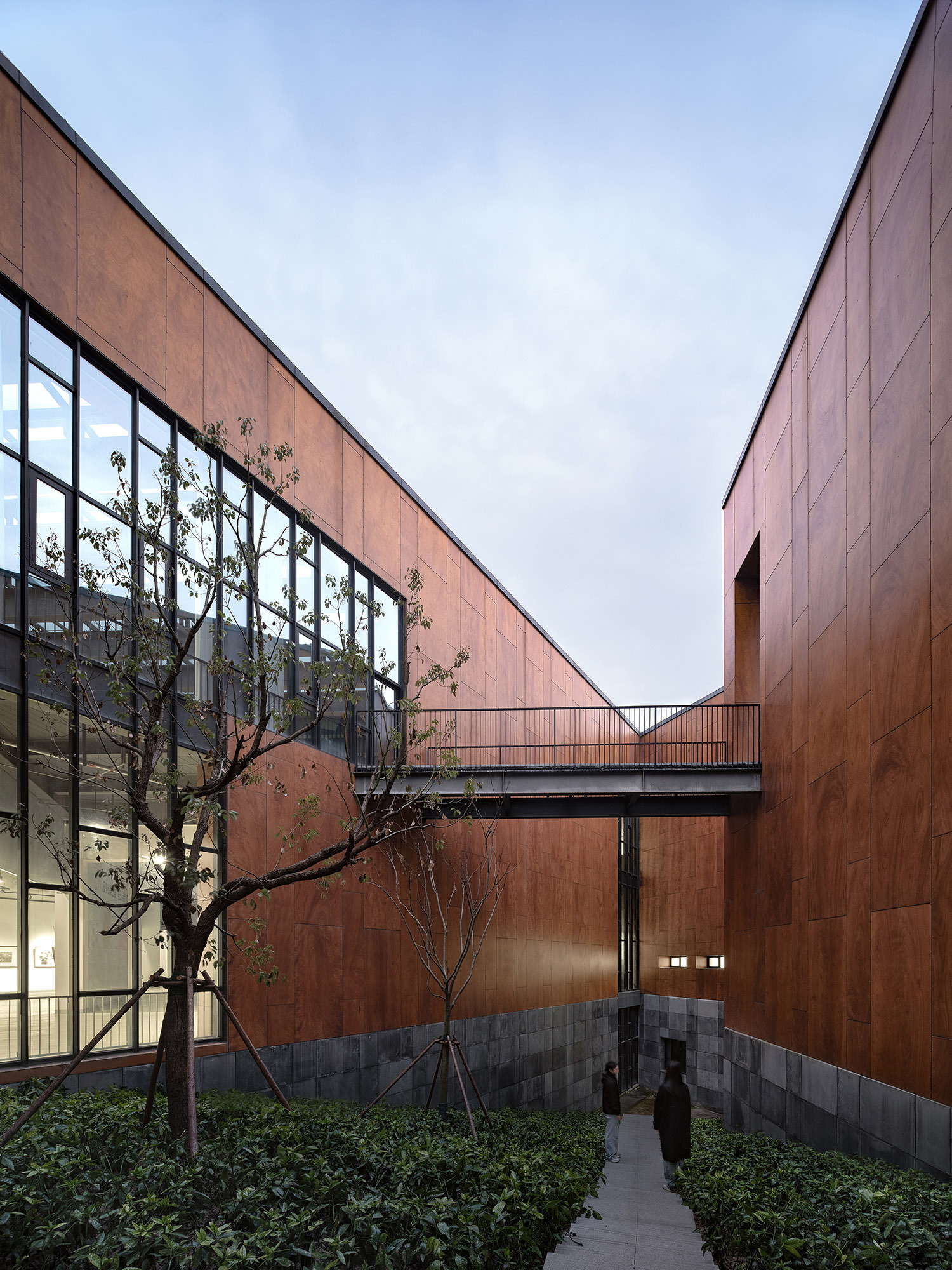 Footbridge spanning a narrow courtyard between rust-brown panel walls, with a young tree and two visitors on the path at dusk