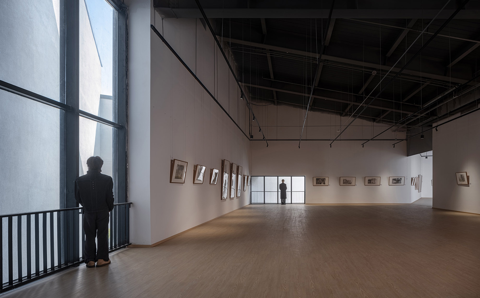Gallery hall with framed drawings along a white wall, exposed dark ceiling trusses and a visitor standing at the tall window