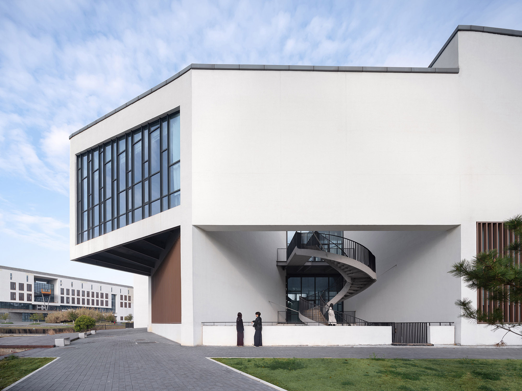 White cantilevered volume above an open ground level with a spiral stair, seen across a paved path under scattered clouds