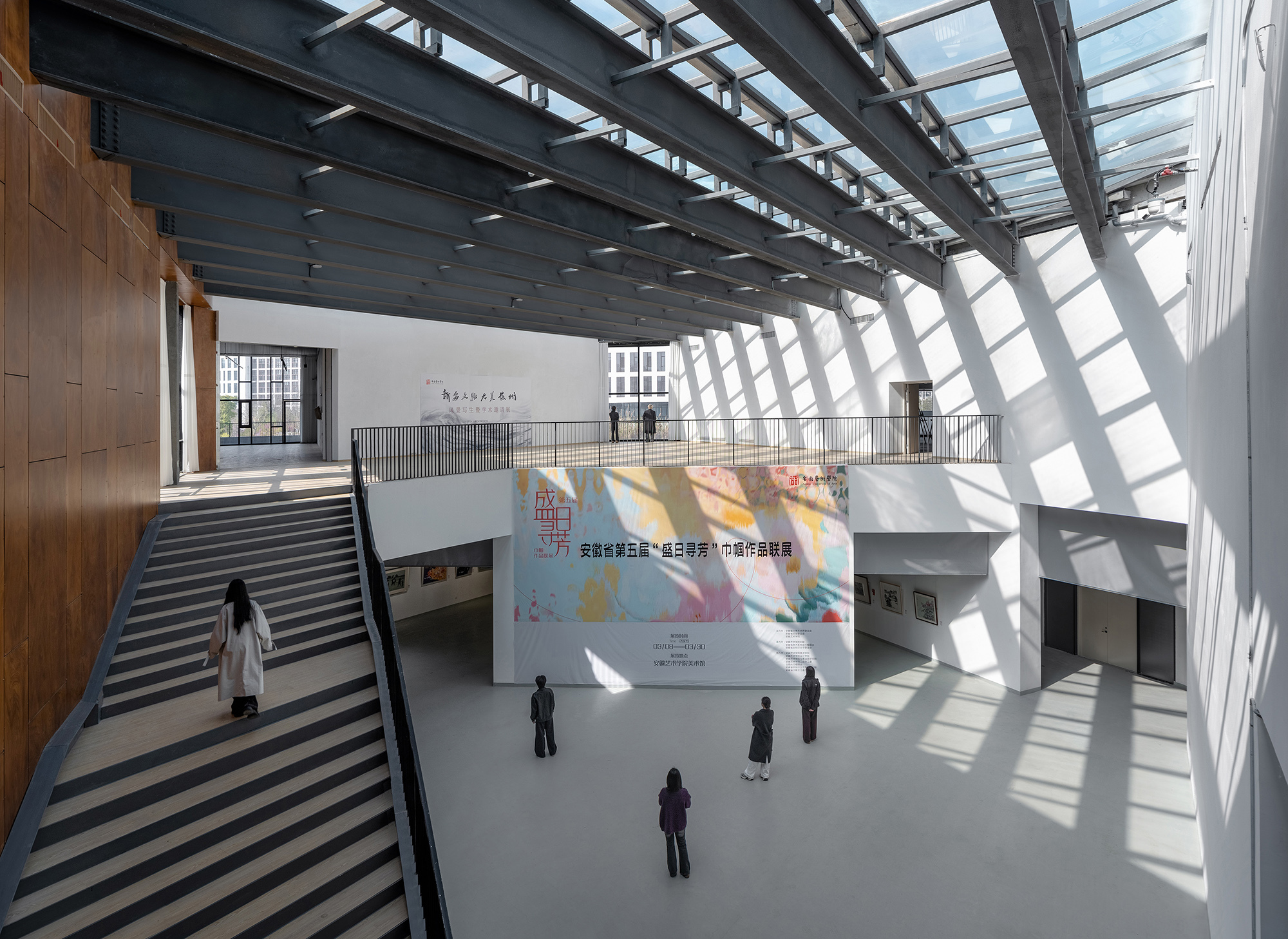 Atrium with a striped stair, steel roof beams and a glazed skylight casting sharp diagonal sunlight across the white walls and visitors