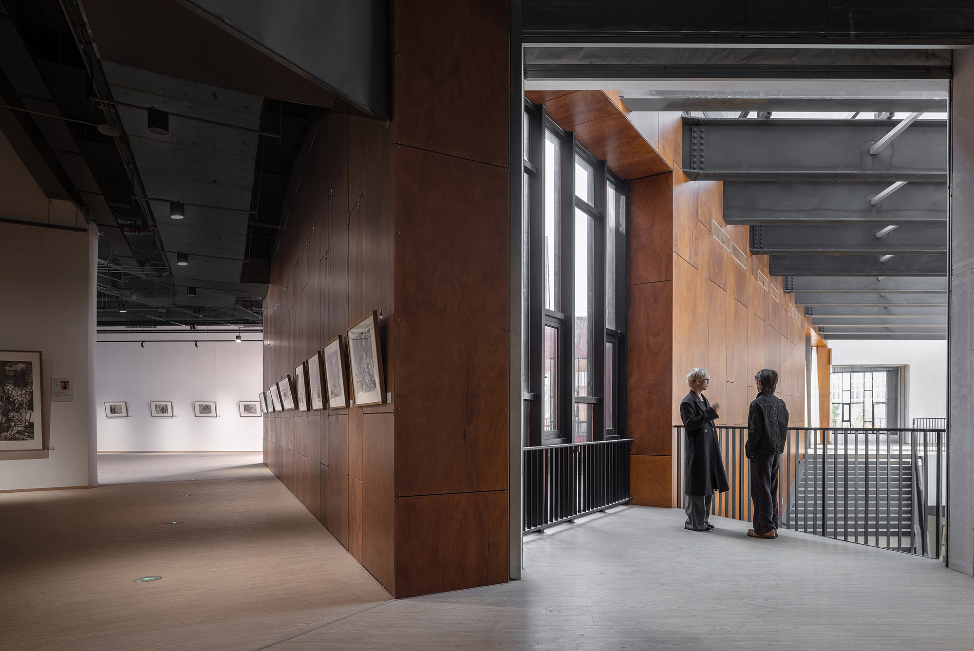 Upper gallery corridor with a wood-clad wall hung with framed prints, and two people talking beside a black steel railing