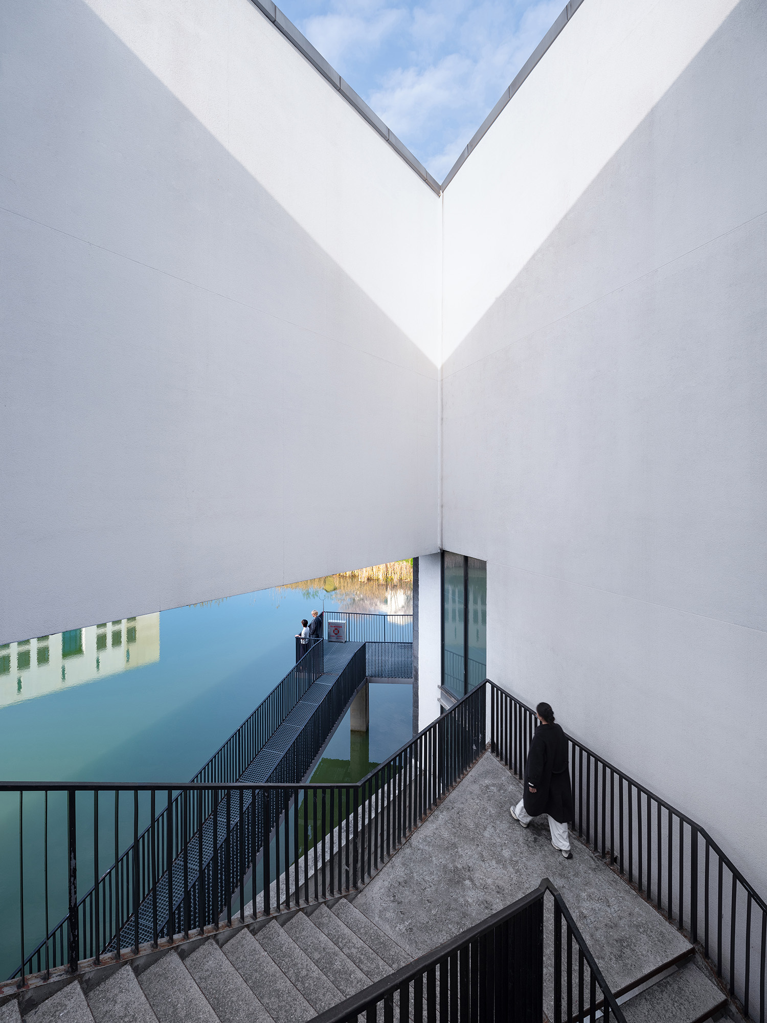 White walls forming a V-shaped sky opening above a concrete stair and black railings, with a person descending toward the water