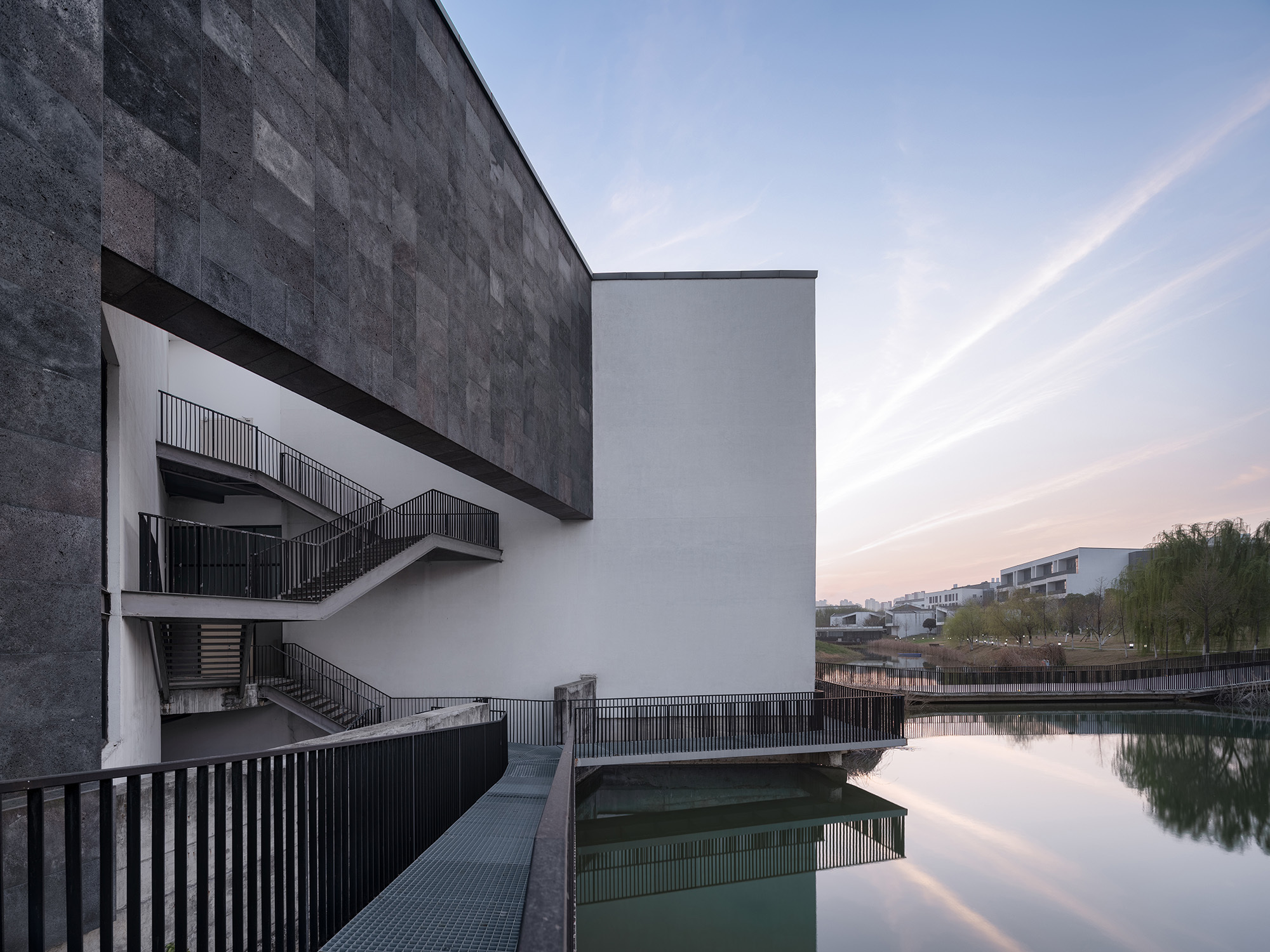 Dark stone-clad volume above a white wall with steel stairs and a grated walkway extending over the pond at dusk
