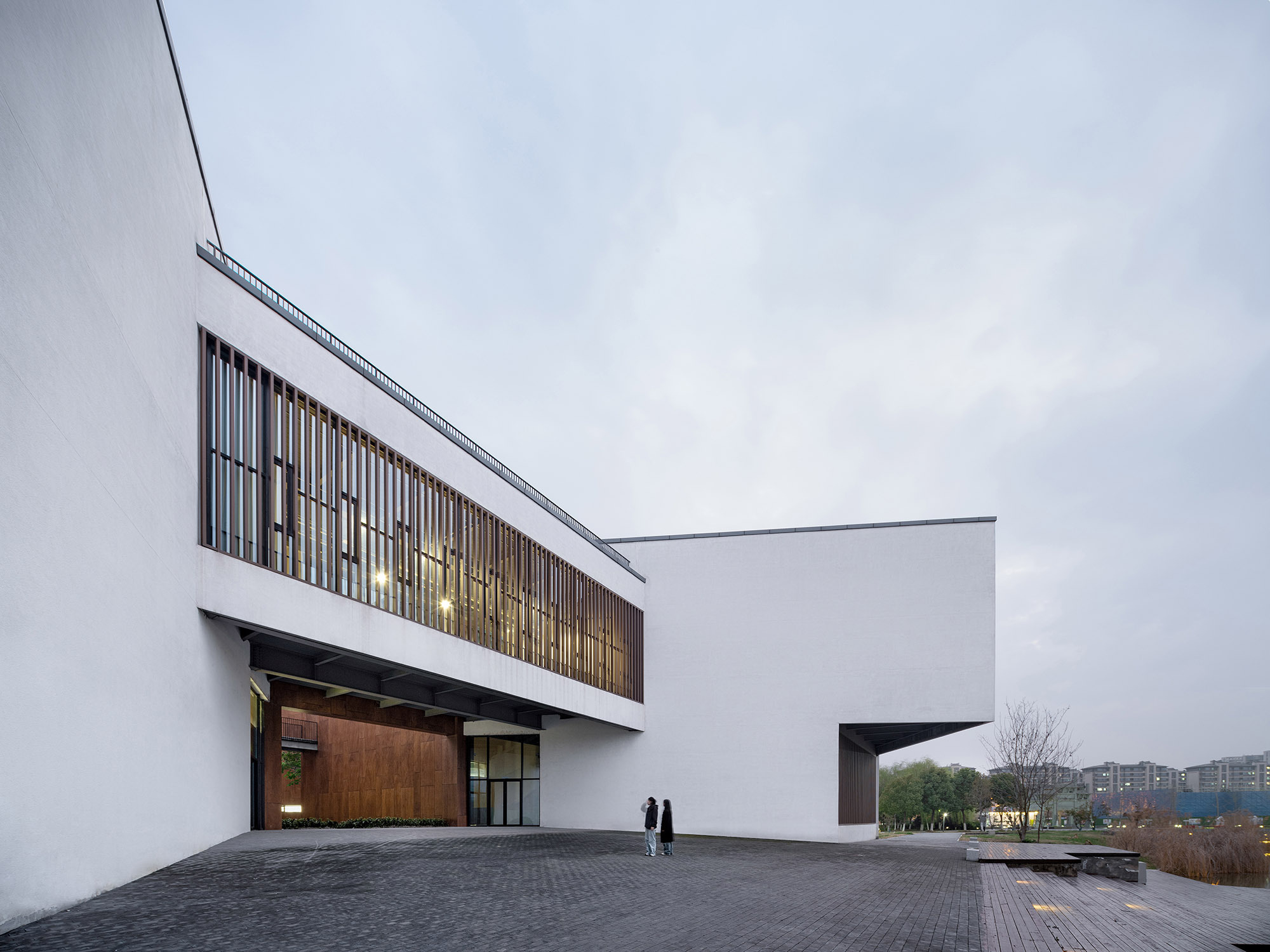 White cantilevered volume with timber slat windows and a rust-toned entrance recess, two visitors standing on the paved plaza under overcast sky