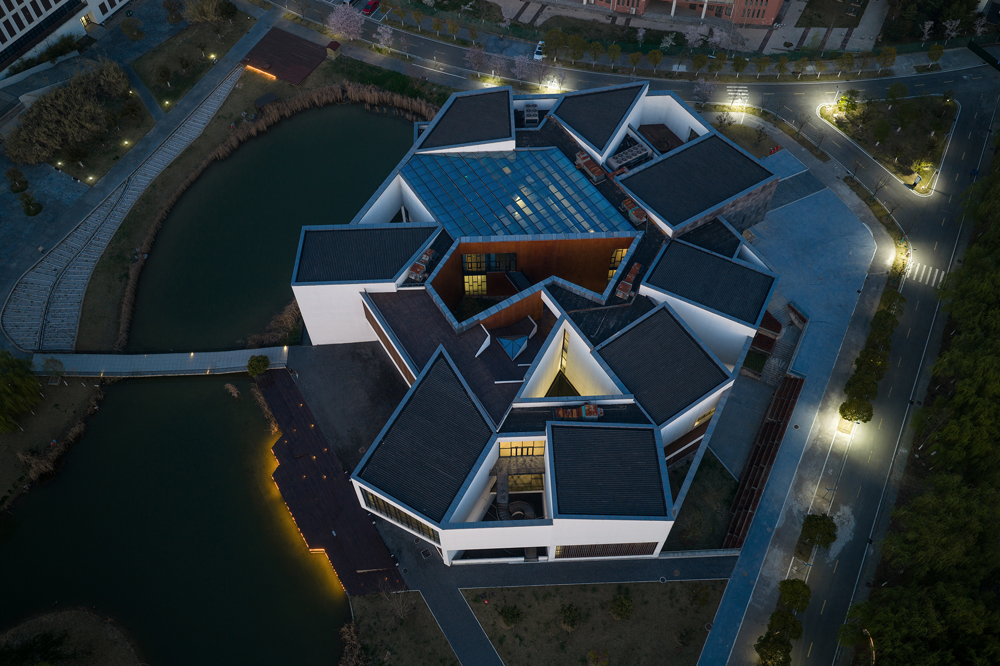 Aerial view at dusk of the pinwheel cluster of pitched dark roofs around a central glazed skylight, beside ponds and a lit road