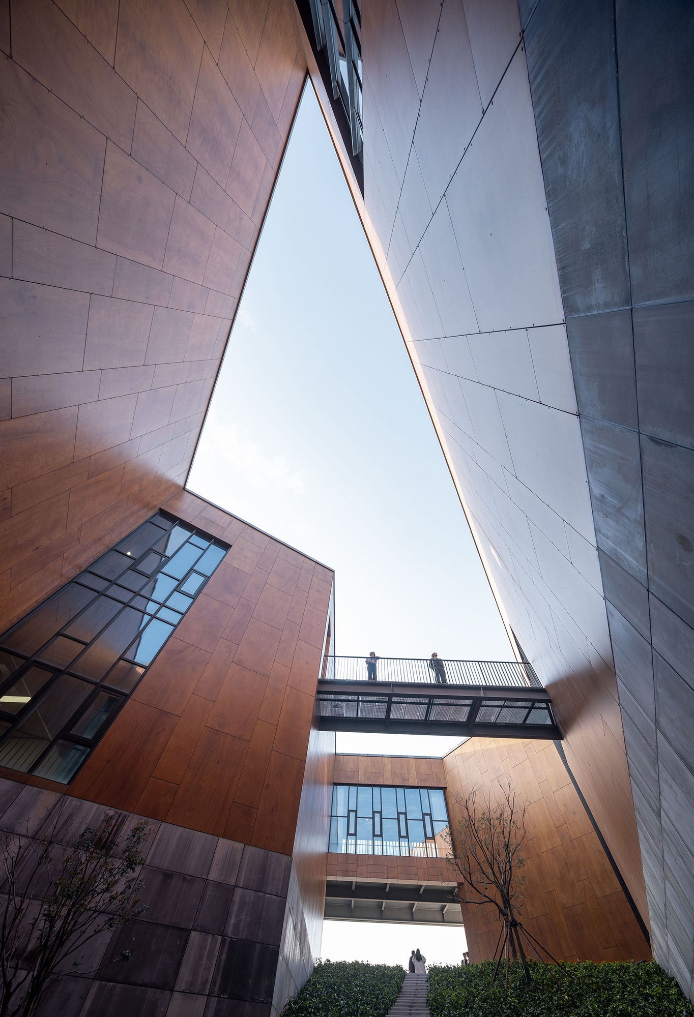 Looking up through a narrow gap between rust-toned panel walls and grey stone, with a glazed bridge and two people crossing