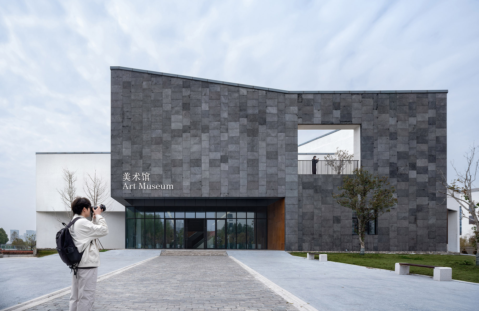 Grey stone facade with a square terrace opening and glazed entrance, while a photographer with a backpack aims a camera in front