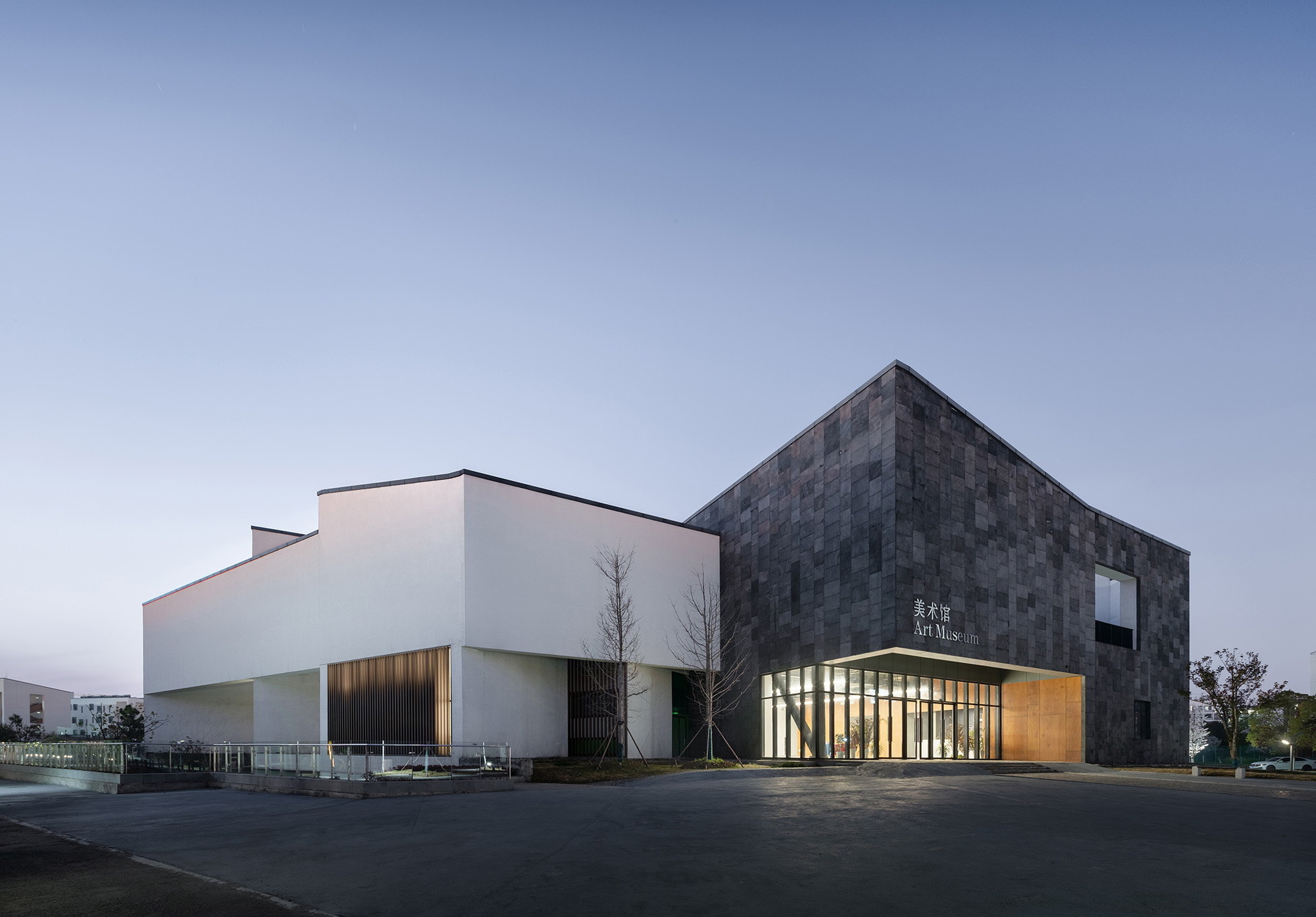 Corner view of the white rendered volume beside the dark slate-clad block, glazed entrance lit at dusk