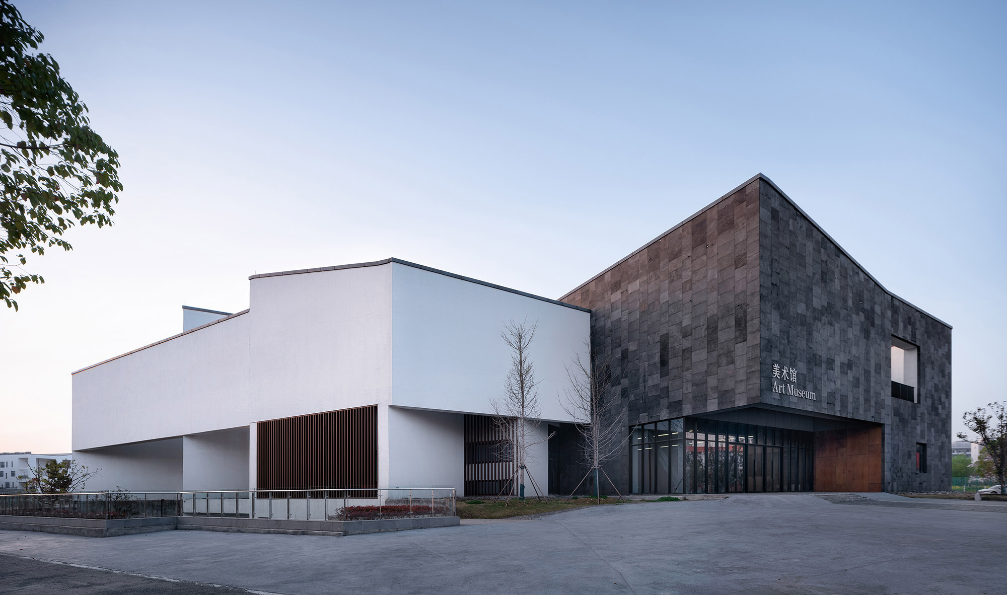 Facade of white walls, timber slats and slate cladding with a glazed entry, seen across an empty plaza at twilight