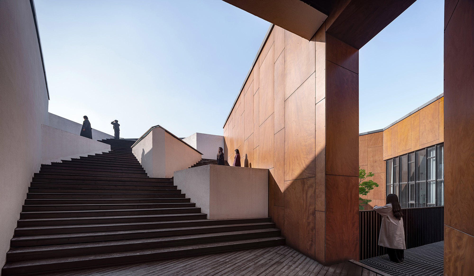 Wide dark timber staircase rising between white walls and plywood-clad volumes, visitors standing on the landings under clear sky