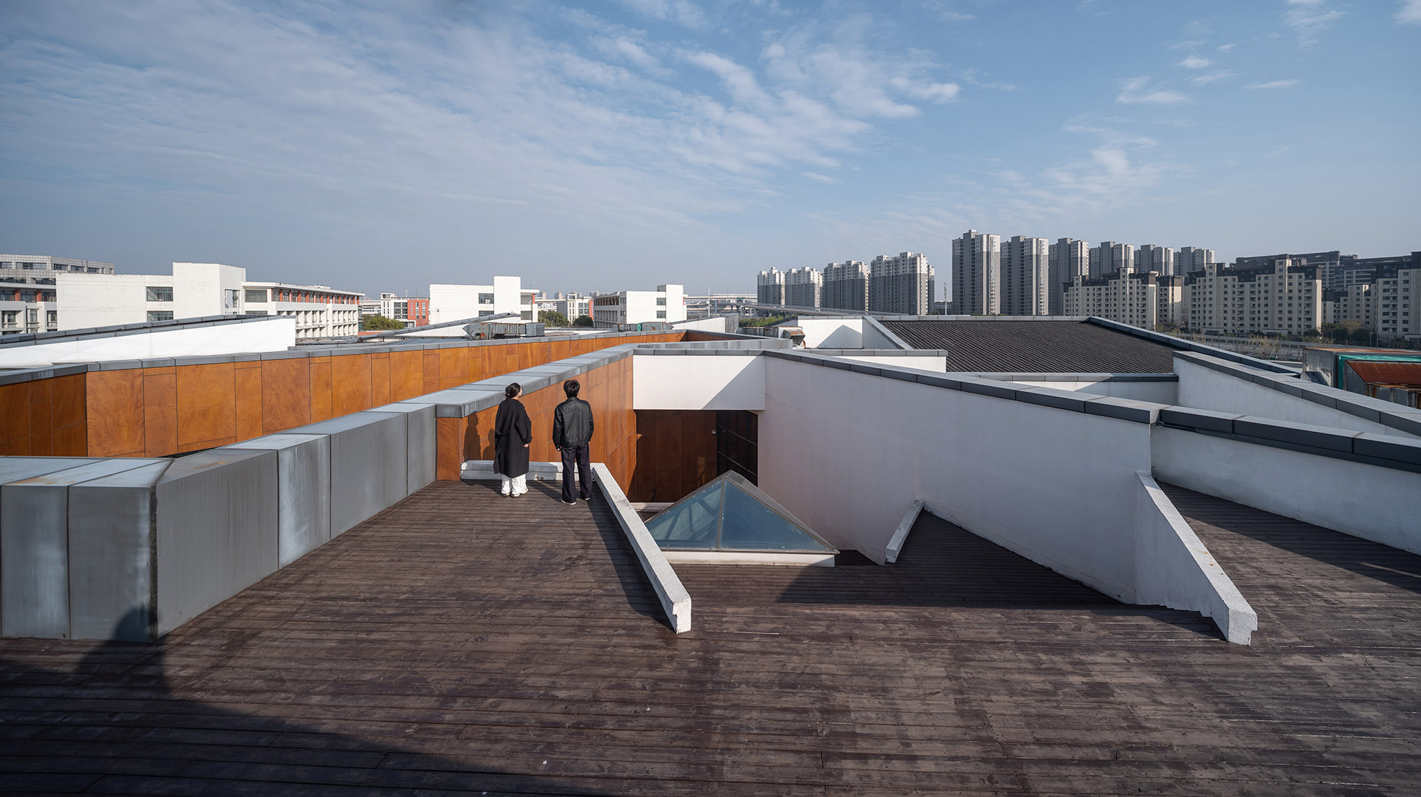 Timber roof terrace with a skylight and rust-toned cladding, two visitors standing before a distant skyline of apartment towers