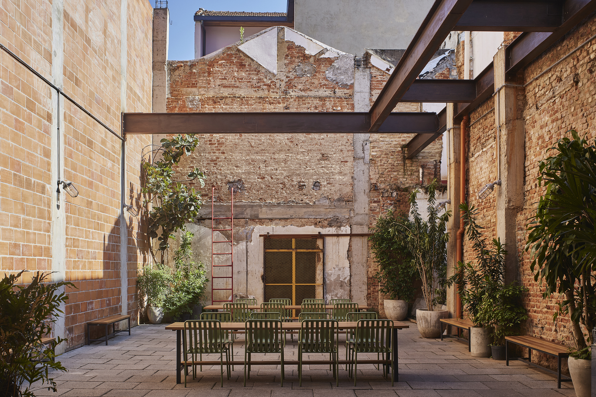Open-air brick courtyard with a long timber table, green metal chairs and potted plants under steel beams in strong sunlight