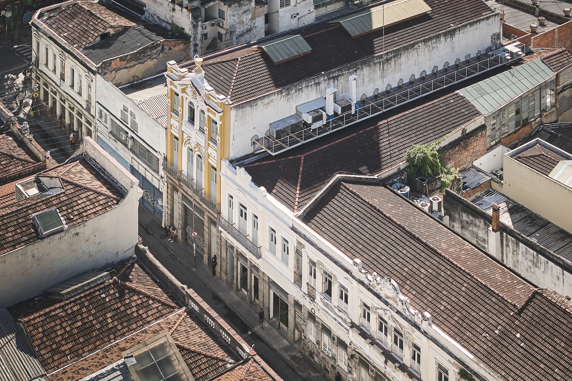 Aerial view of terracotta tile roofs and pale painted facades, with a yellow ornate building and rooftop air conditioning units in sun