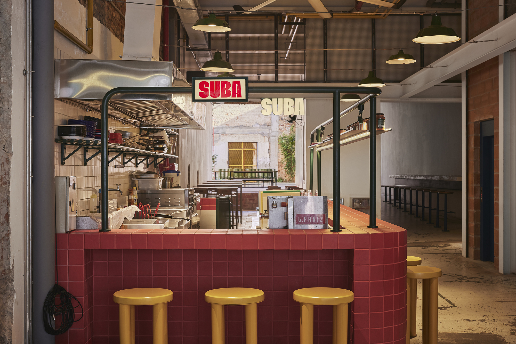 Red tiled counter with yellow stools and a lit sign, looking through to a sunlit brick courtyard beyond