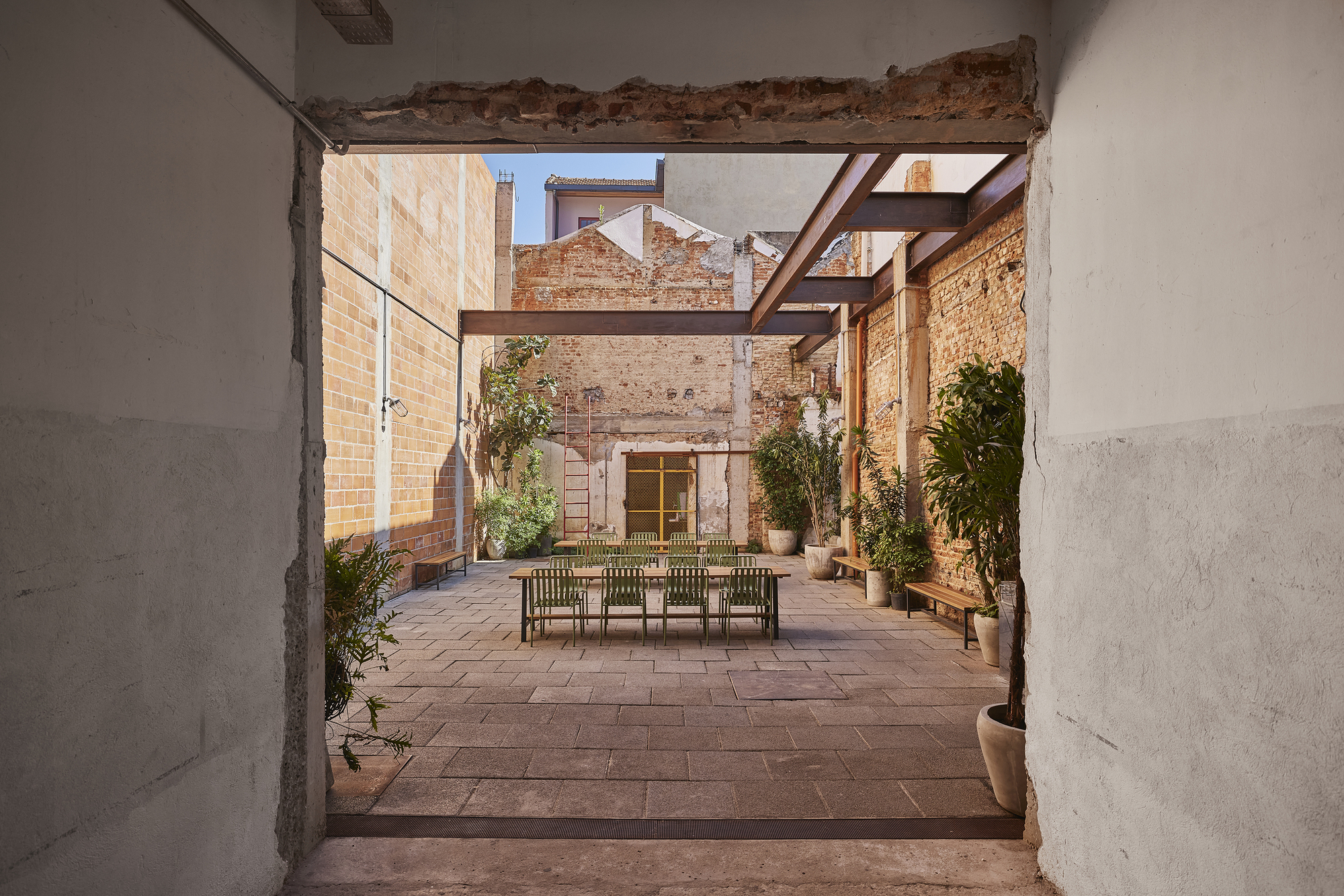 View through a rough plastered opening into a paved courtyard with a dining table, steel beams and climbing plants in sunlight