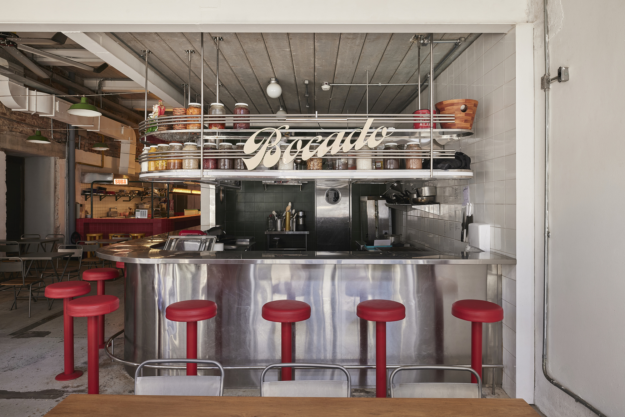 Stainless steel counter with red stools beneath a suspended shelf of jars and script lettering, with green pendant lamps overhead