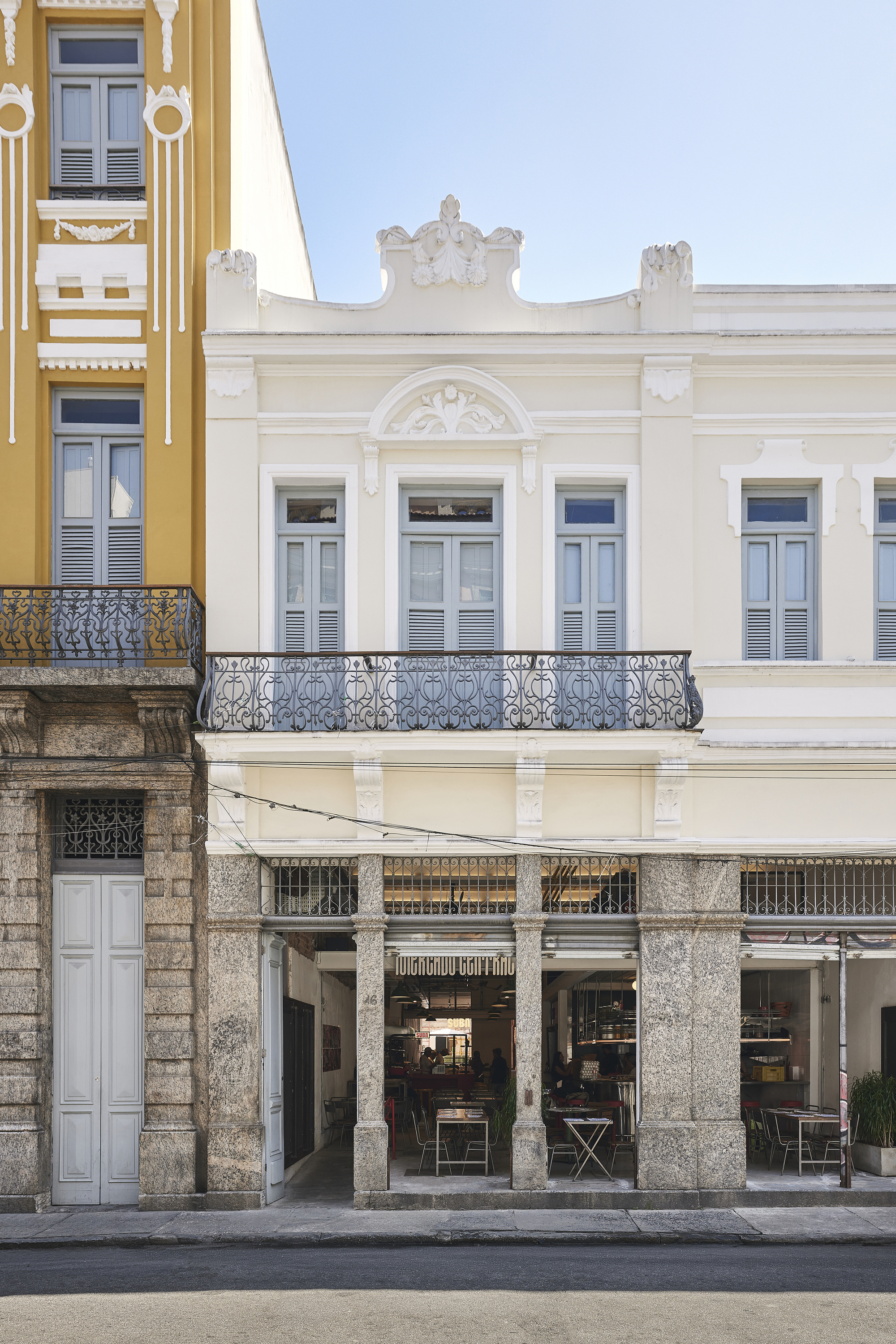 Street facade of a cream building with a wrought iron balcony, blue shutters and granite columns above an open ground floor, beside a yellow neighbor