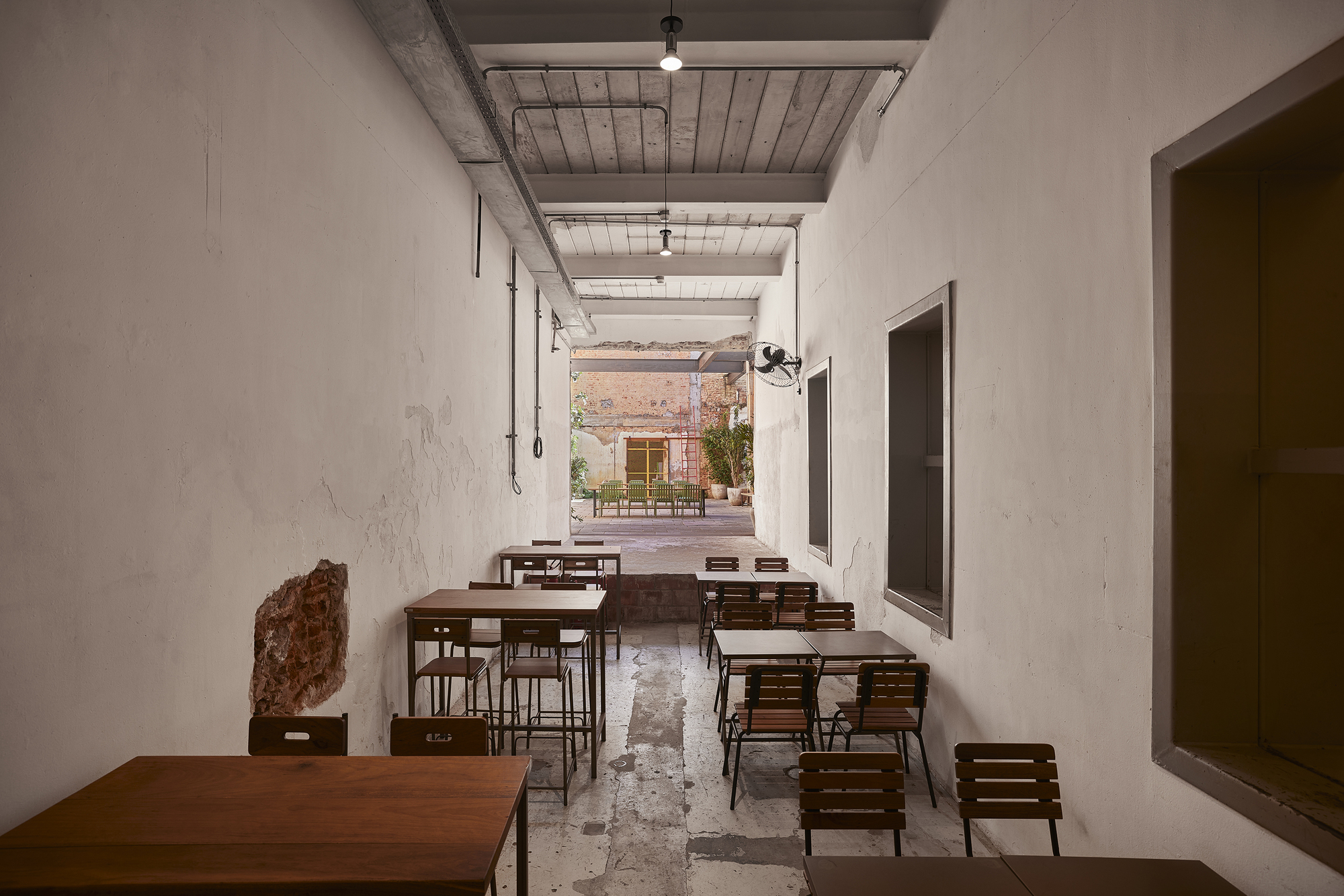 Narrow side hall with wooden tables and slatted chairs leading toward a sunlit rear courtyard with potted plants