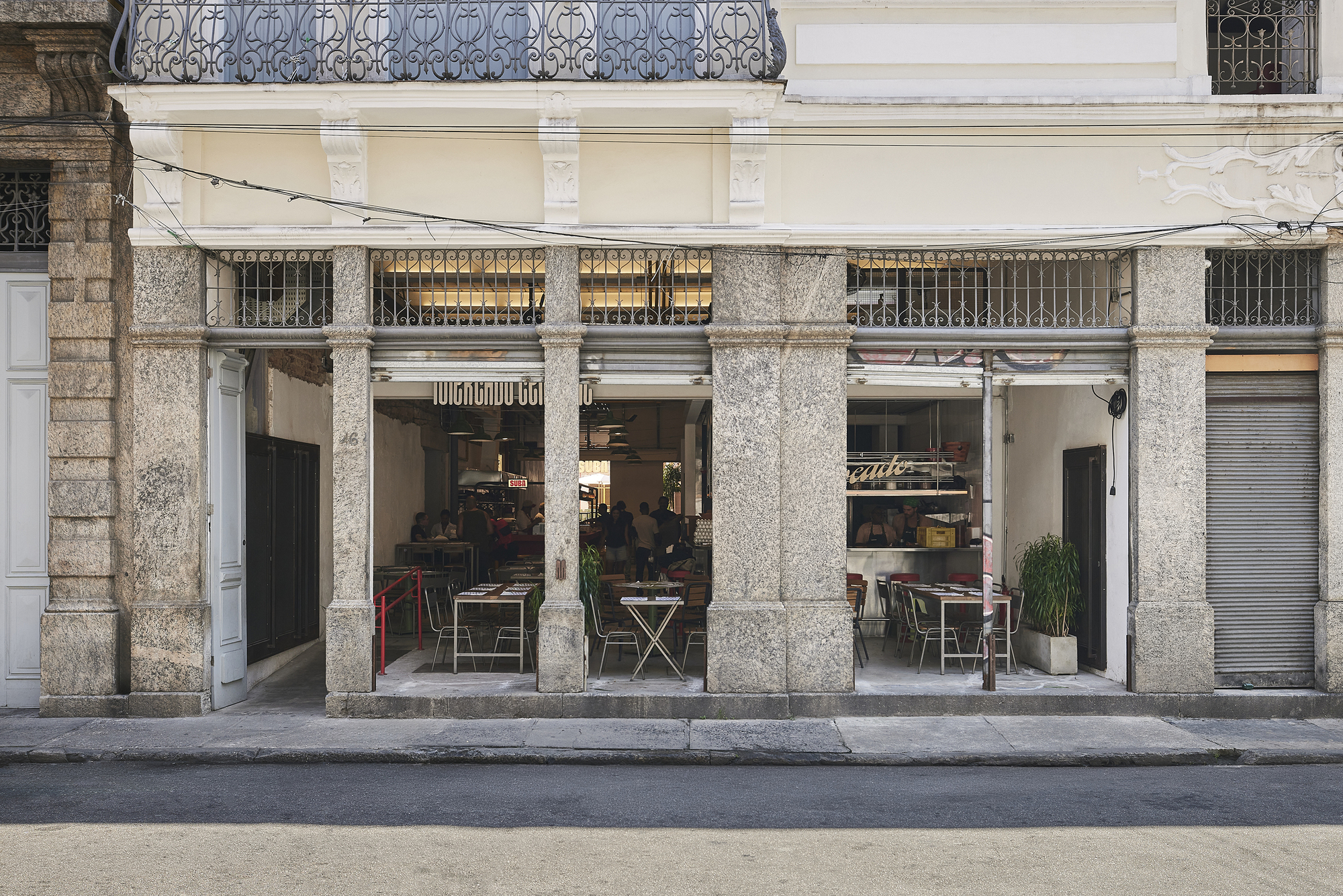 Street facade of granite piers and iron grilles opening onto a restaurant with diners seated inside in daylight