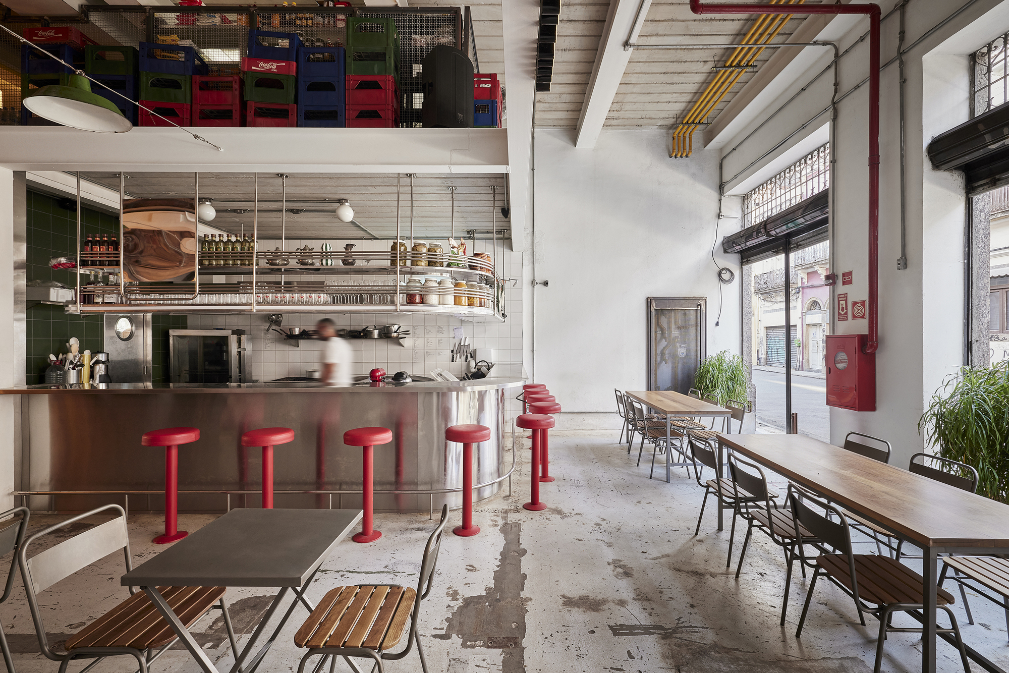 Dining hall with a stainless steel counter, red stools and stacked crates above, a blurred cook moving behind it