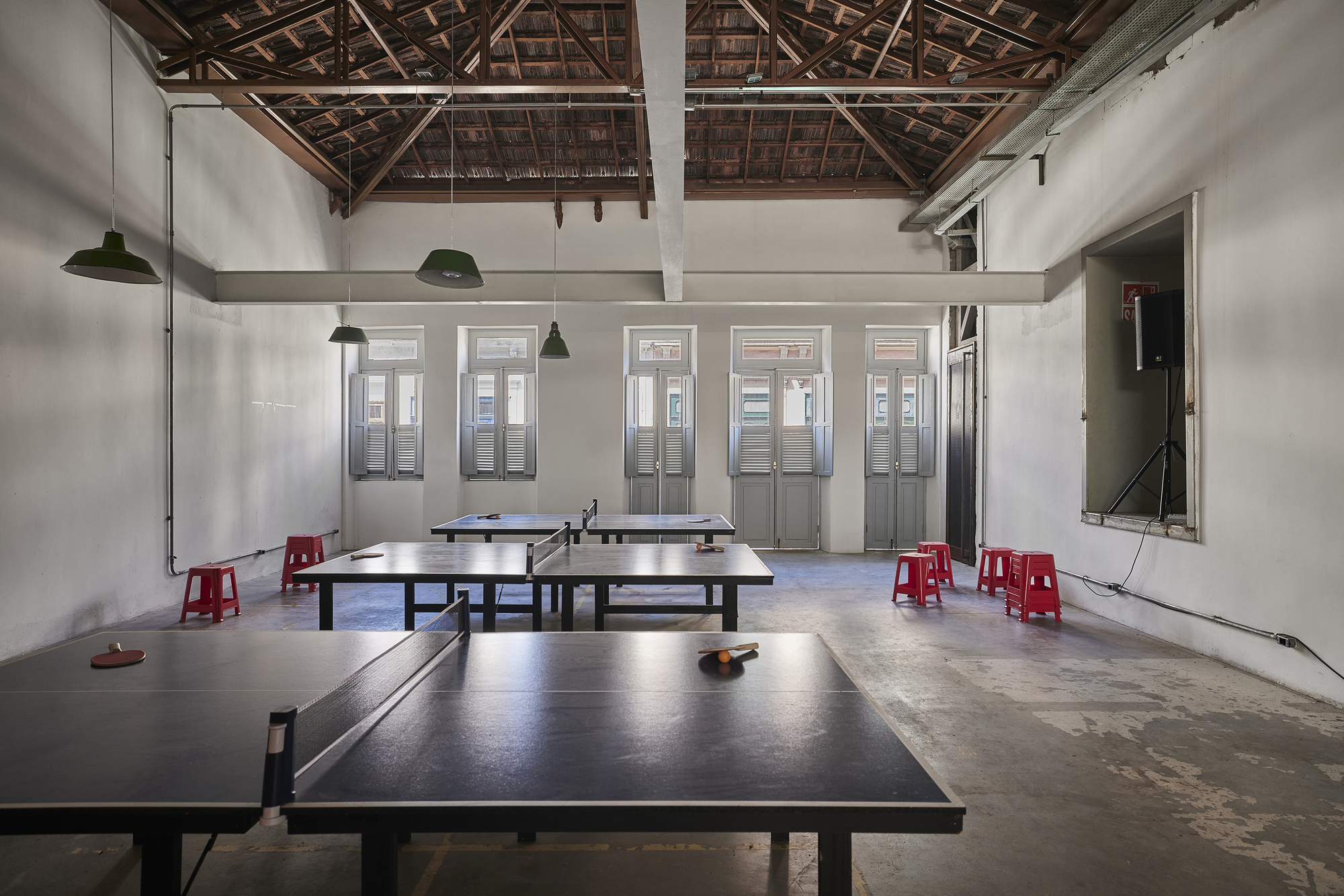 Open room with ping pong tables, red plastic stools and exposed timber roof trusses above shuttered tall doors
