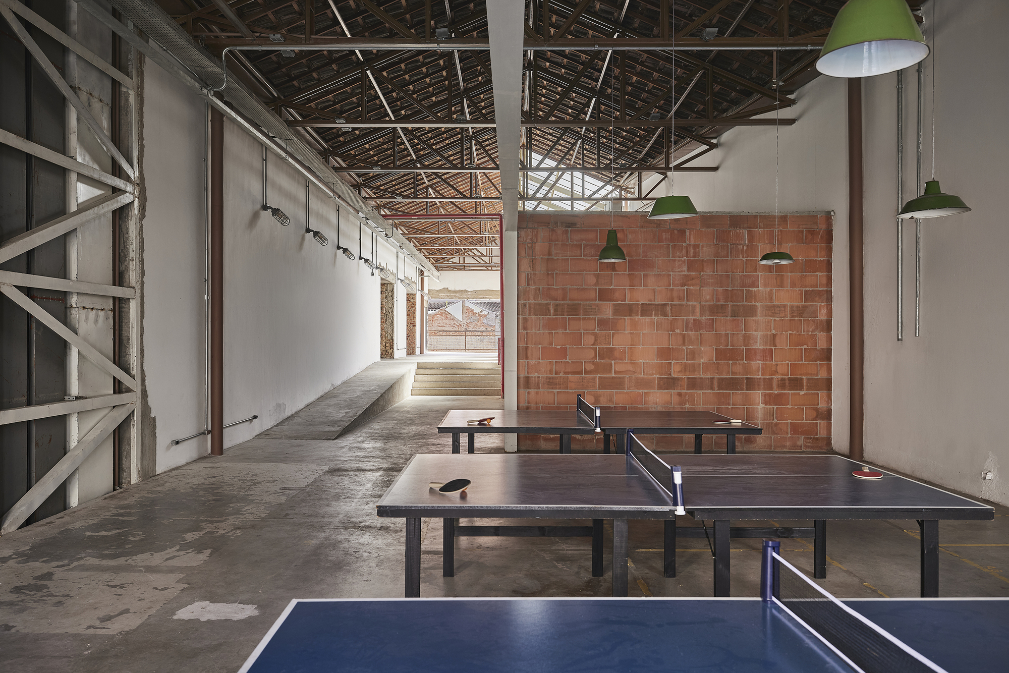 Ping-pong tables in a converted hall with a terracotta block wall, green pendant lamps and exposed roof trusses