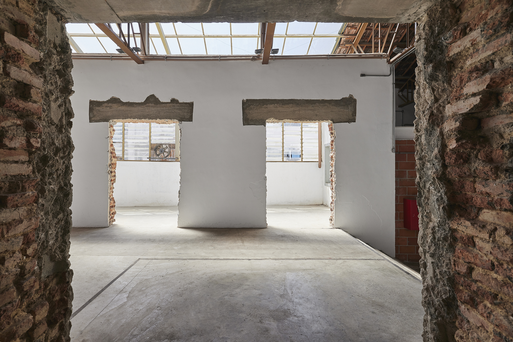 Two cut openings with exposed concrete lintels in a white wall, framed by rough brick edges and skylights above