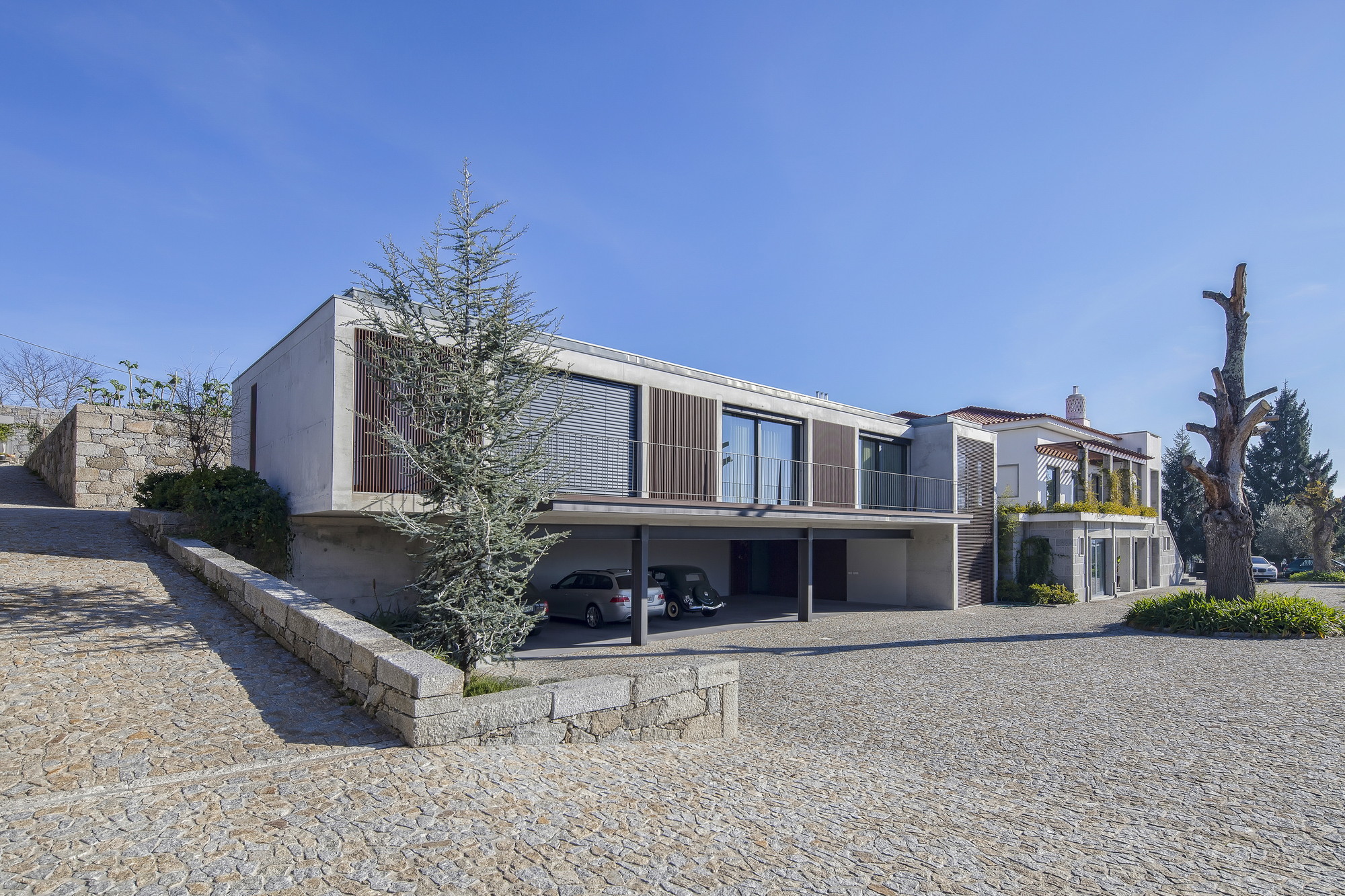 Concrete and timber-shuttered extension raised over a carport, with granite cobbles, a low stone wall and a bare tree stump under clear sky