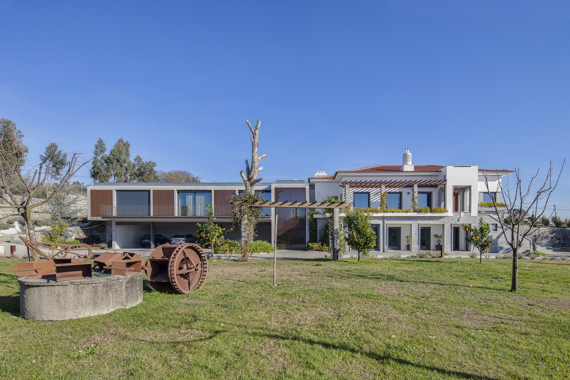 Garden view of the concrete extension beside the white tiled-roof house, with a rusted iron wheel on a round stone base in the foreground