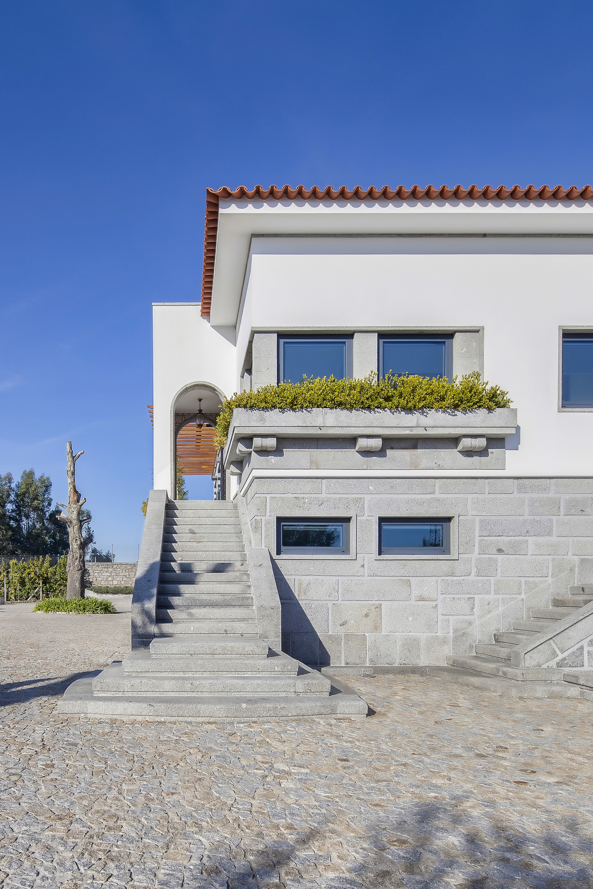 Granite staircase rising to an arched entrance beside a planted stone balcony on the white facade, under a deep blue sky