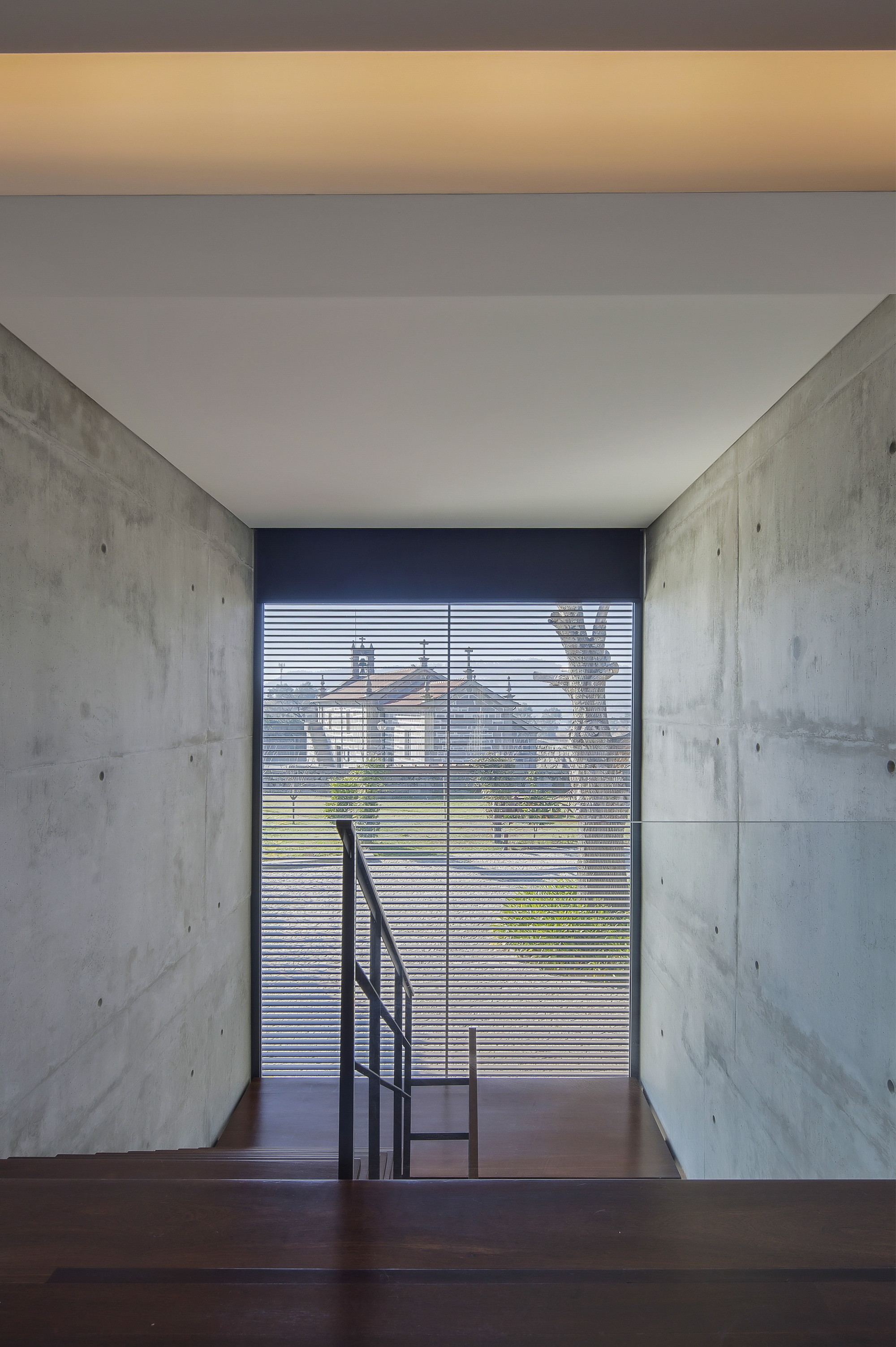 Concrete stairwell with a wooden landing and a slatted glass window framing a distant chapel and a bare tree