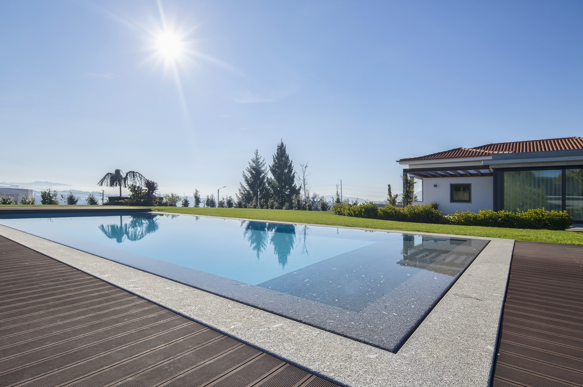 Granite-edged swimming pool with timber decking reflecting trees, the white pavilion and tiled roof beyond, and the sun flaring above