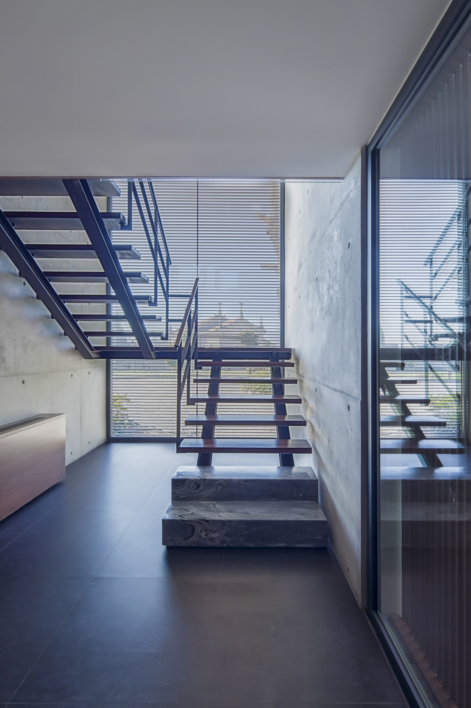 Open-riser staircase with timber treads and steel stringers beside a board-marked concrete wall, lit through a louvered glass window