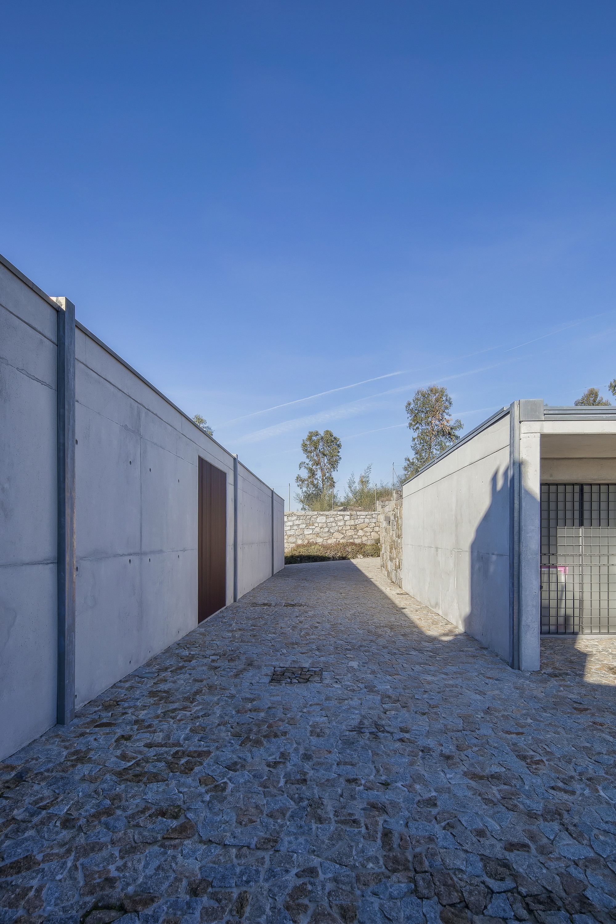 Narrow cobbled passage between board-marked concrete walls, one with a dark timber door, under a clear blue sky