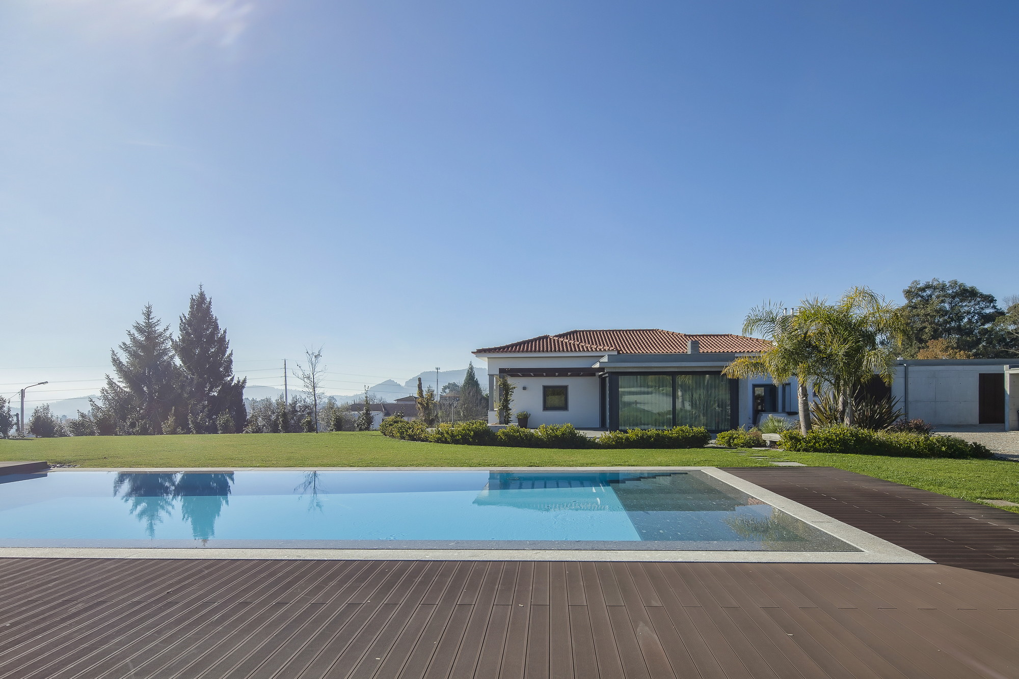 Rectangular pool and composite timber deck in front of a single-storey house with a terracotta roof, palms and lawn
