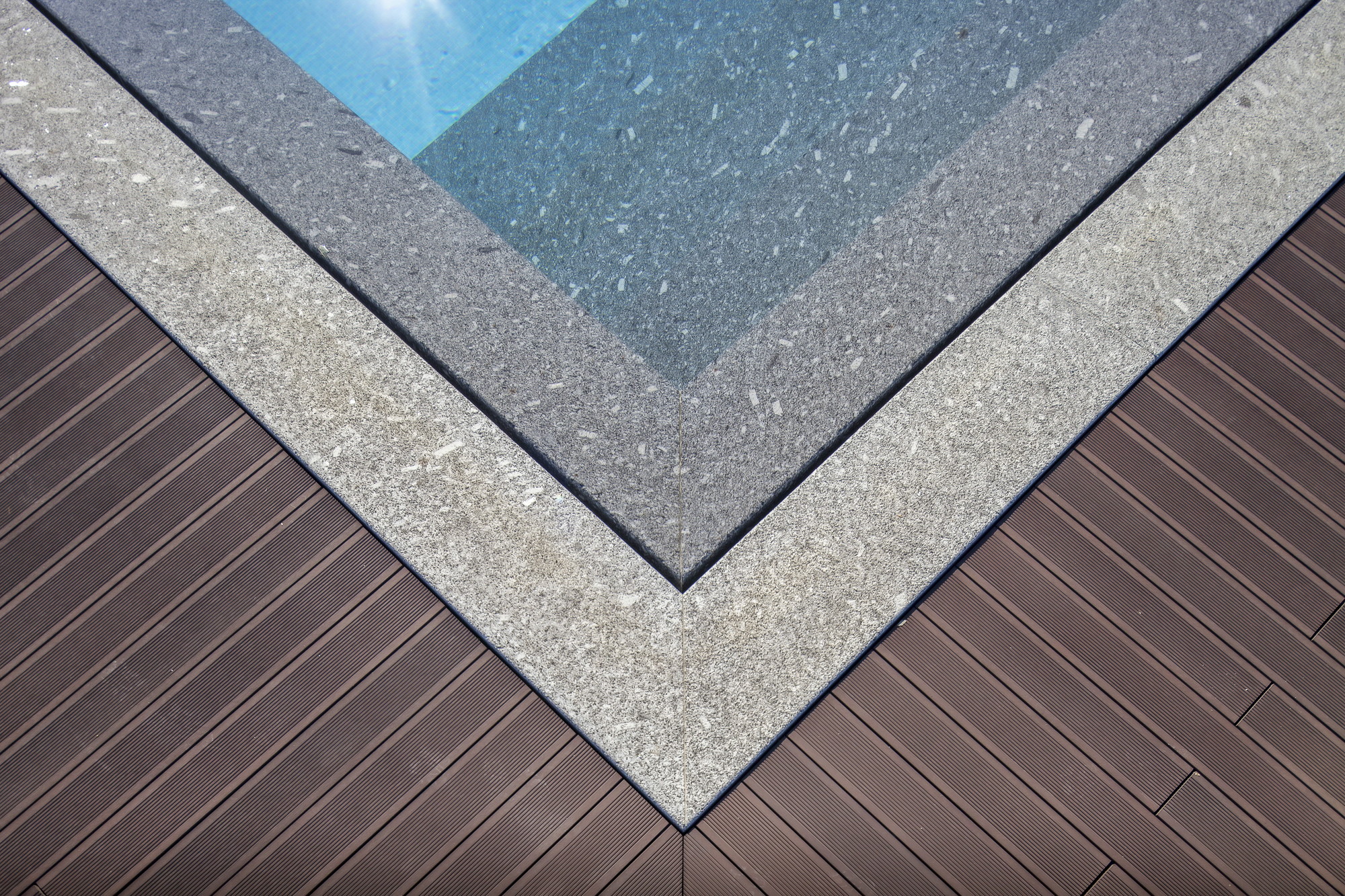 Close-up of a pool corner where speckled granite coping meets dark grooved composite decking in bright sun