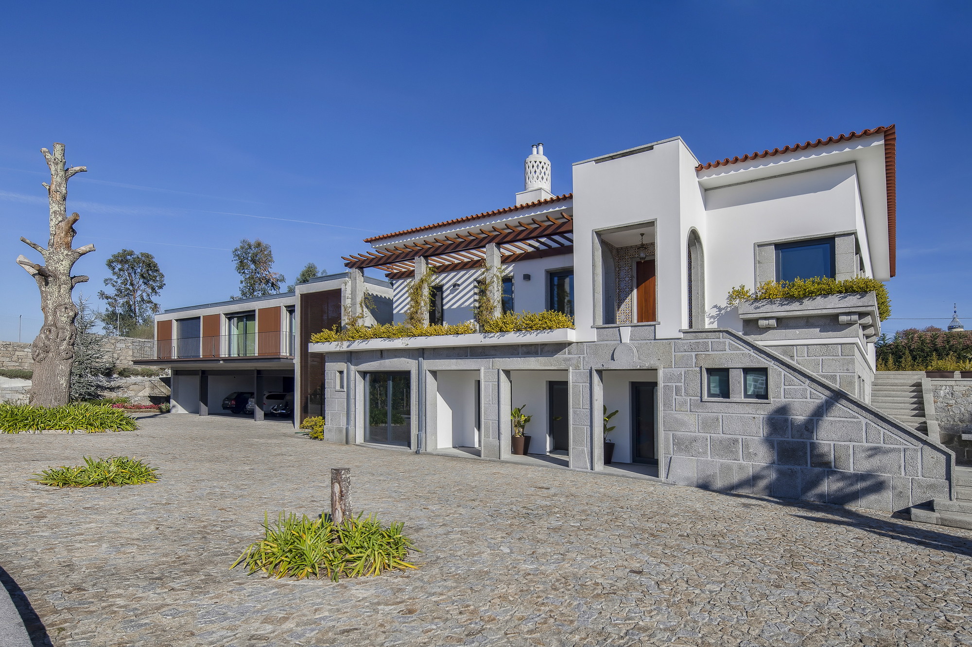 Granite-based house with white upper volumes, a timber pergola and planted terraces above a cobbled forecourt in strong sunlight