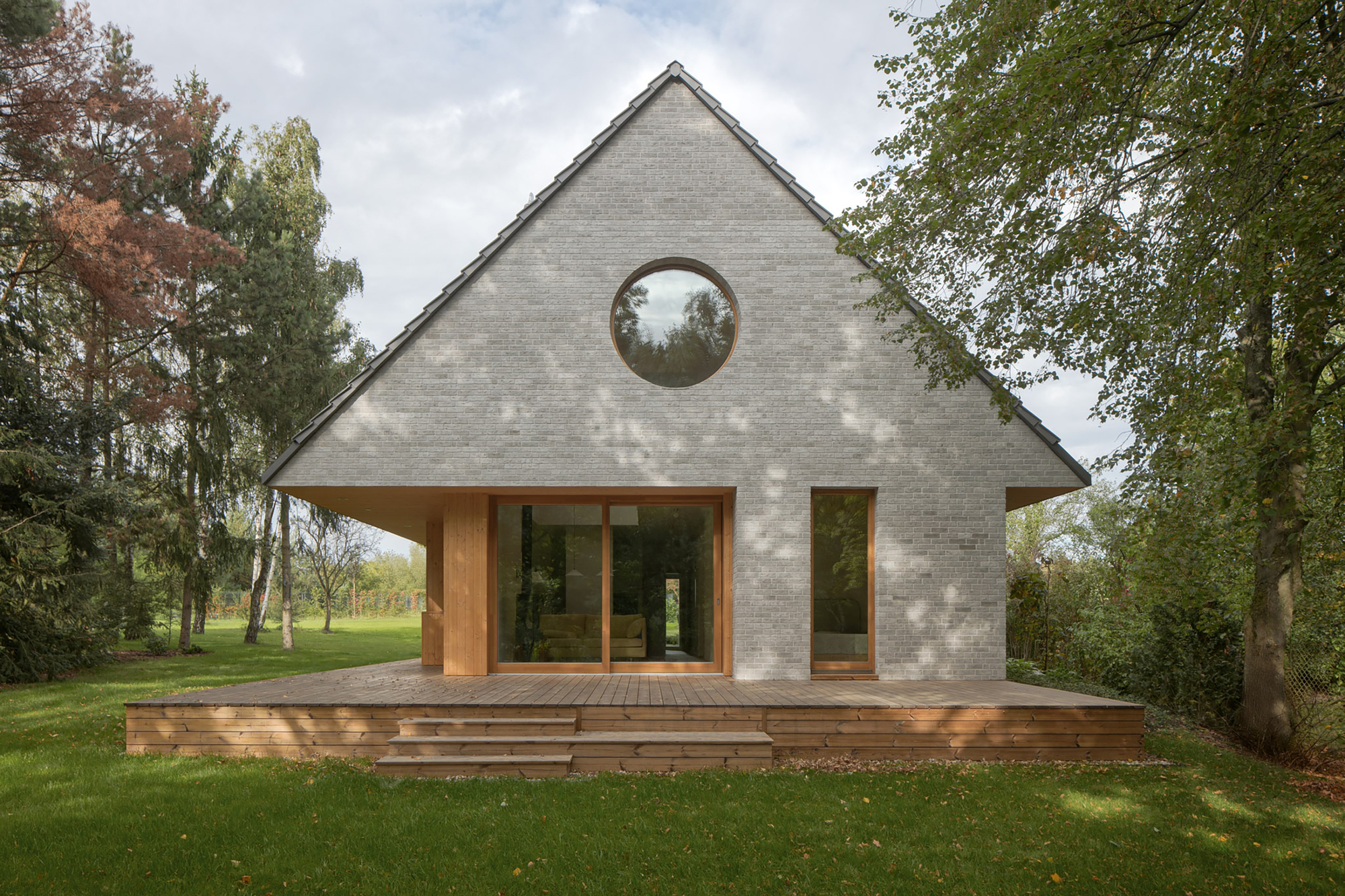 Front gable of pale grey brick with a round window and timber-framed glazing above a low wooden deck, dappled by tree shadows