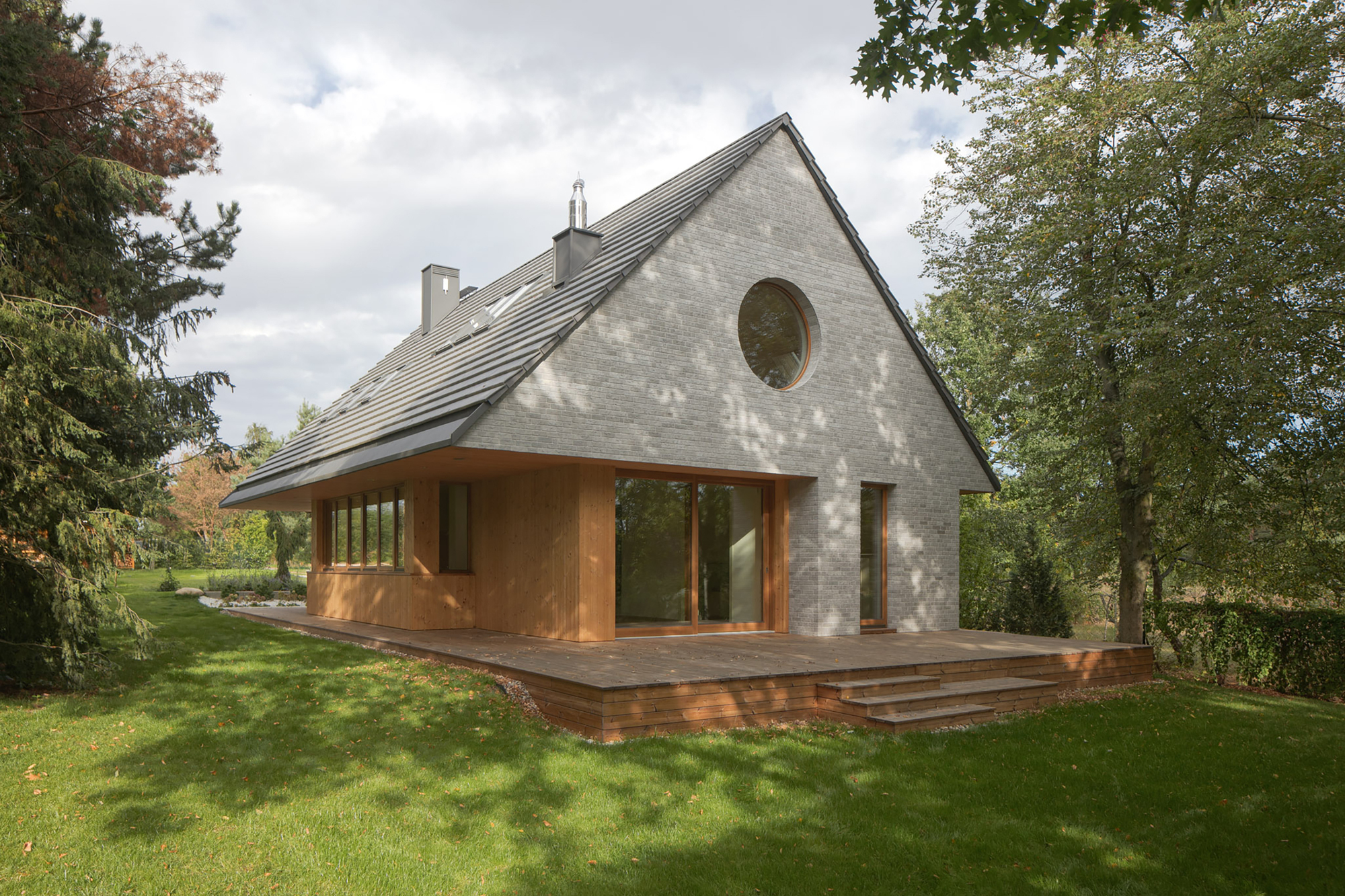 Corner view of the brick gable, slate roof and timber-clad porch beside a wooden deck under a cloudy sky