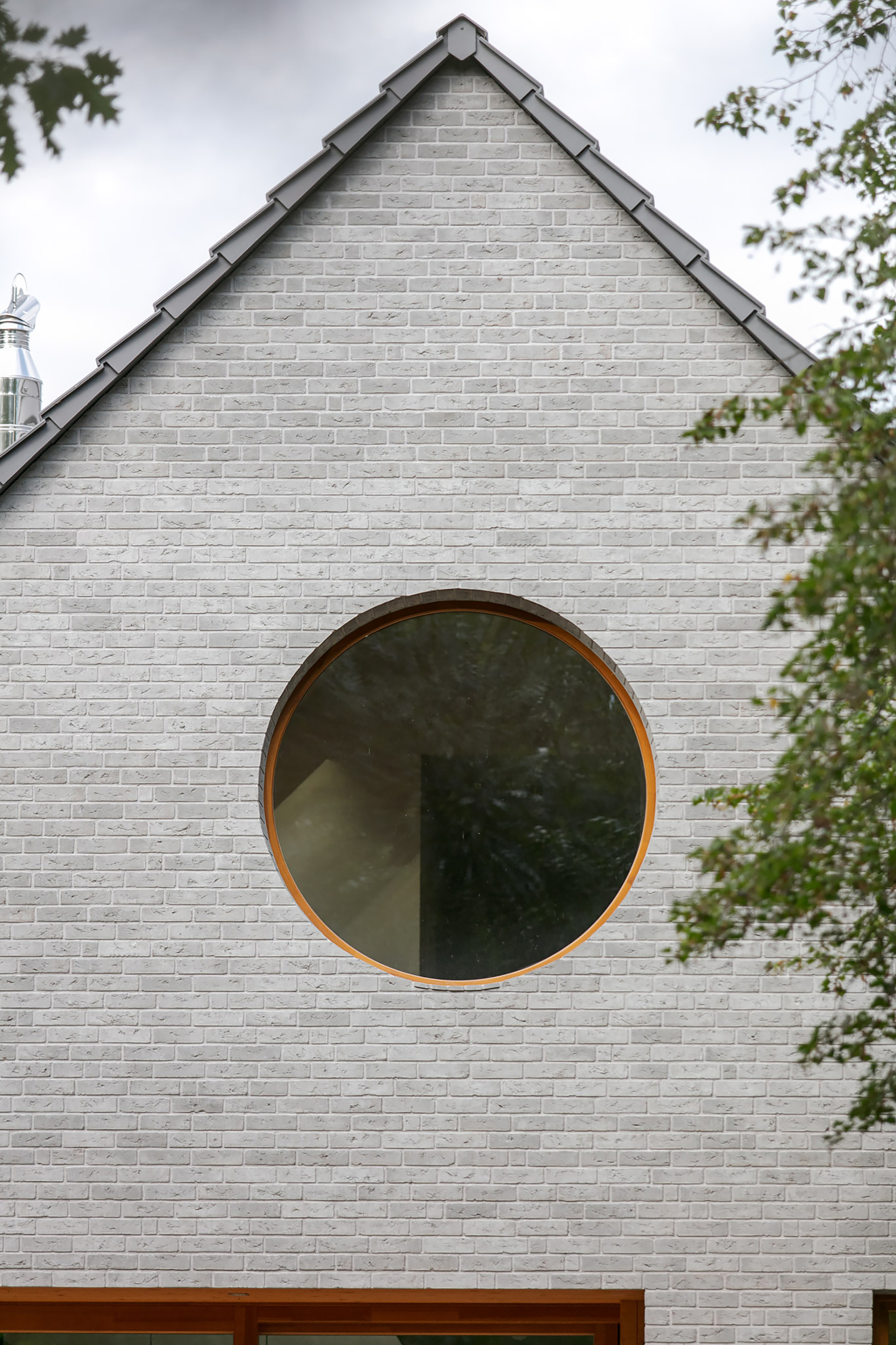 Close-up of the circular timber-framed window set in grey brick below the roof edge, with leaves at the right