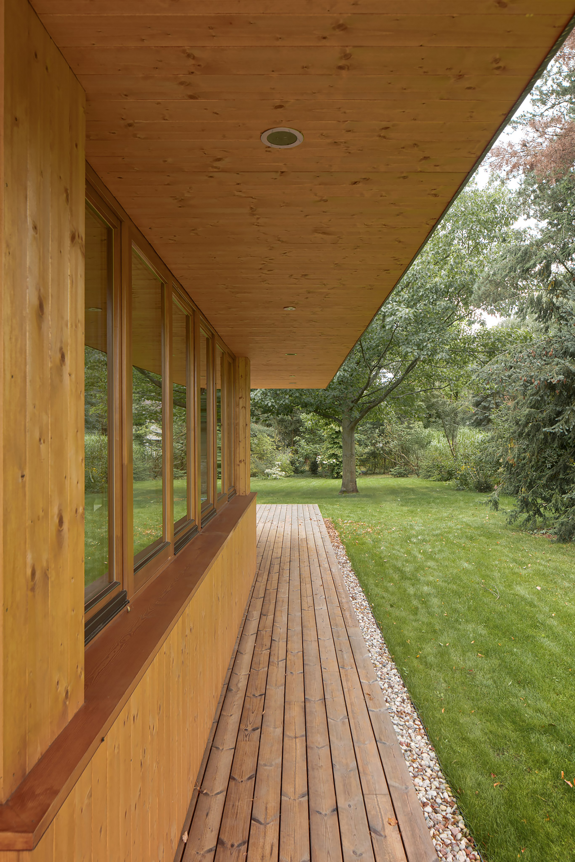 Covered timber porch with a row of narrow windows, plank decking and a gravel strip beside a green lawn