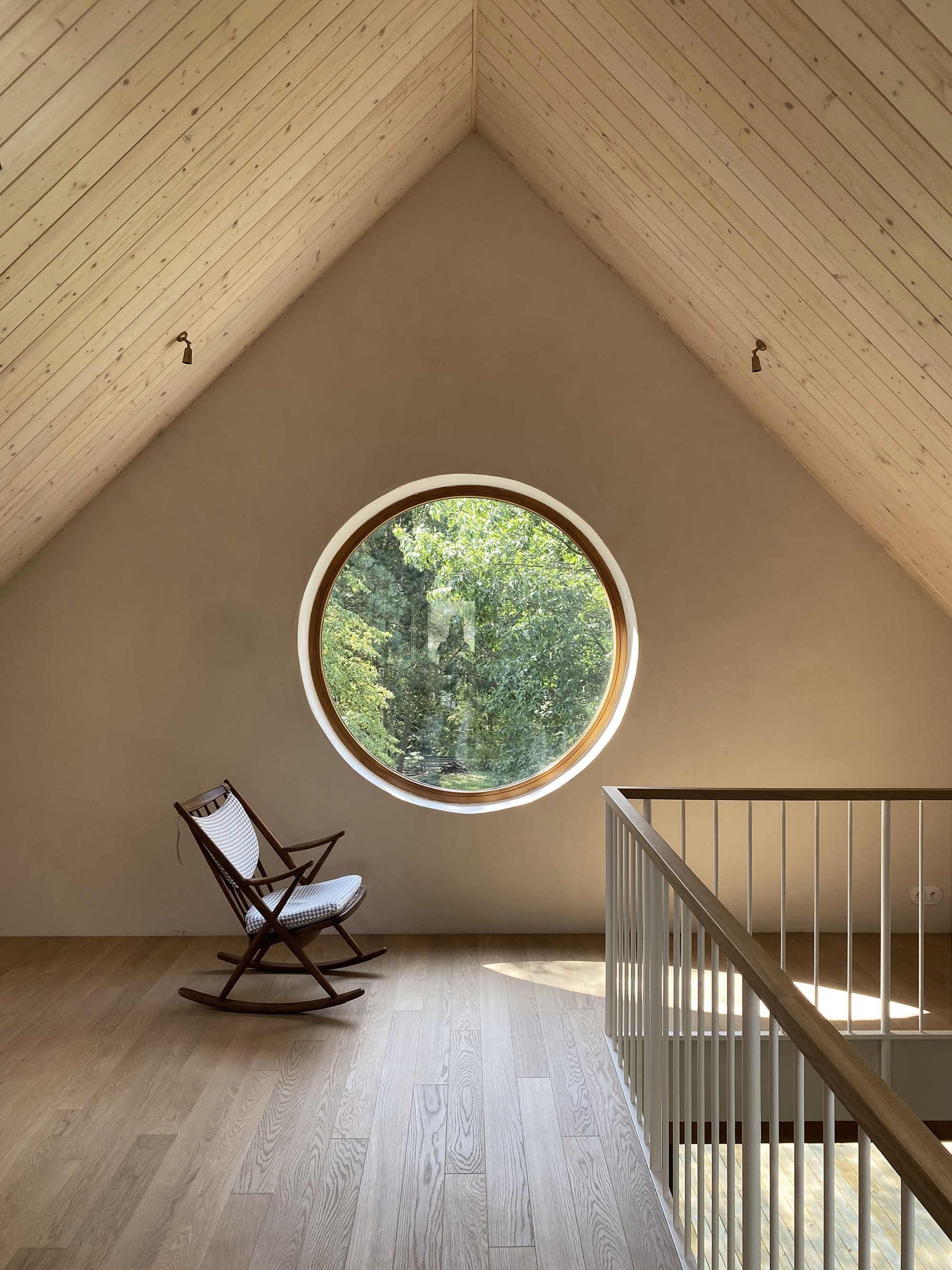 Upper floor under a pitched timber ceiling with a round window, a rocking chair and a white balustrade in sunlight