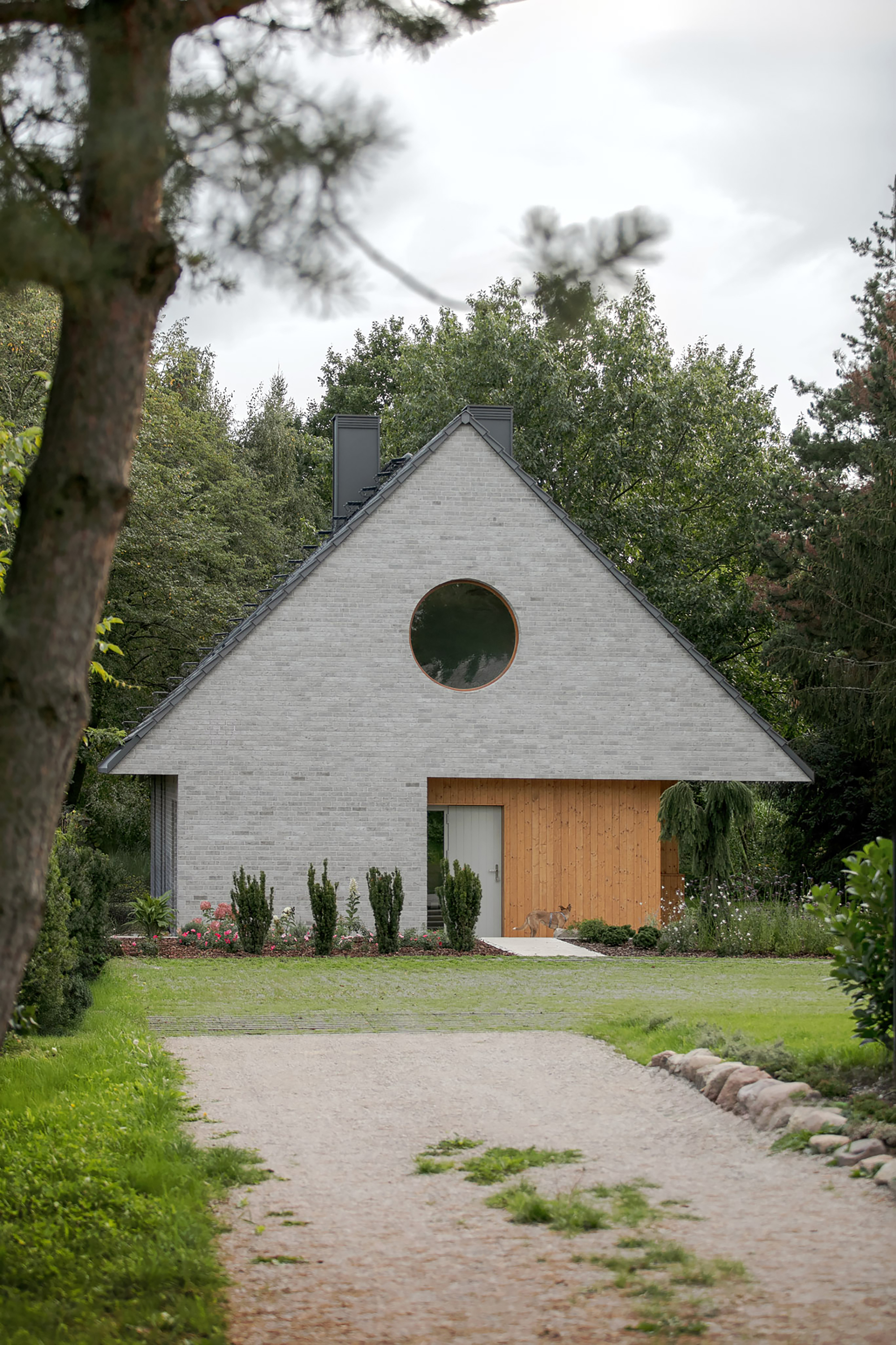 Gabled grey brick facade with a round window and timber entrance bay, seen across a gravel drive under overcast sky