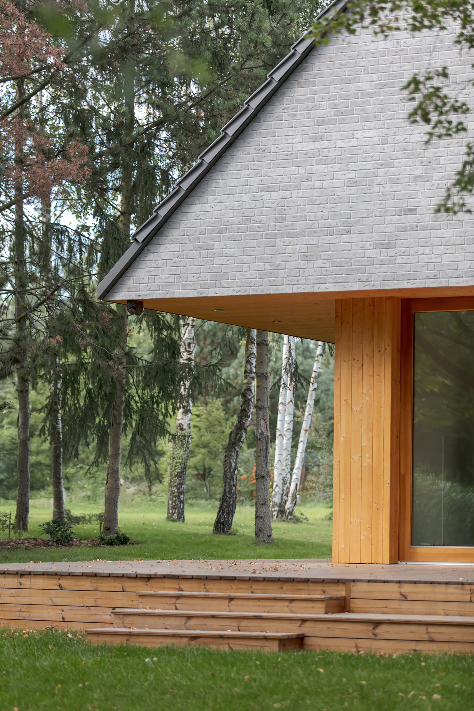 Corner of the grey brick gable above a larch-clad wall and low timber deck steps, with birch trunks behind