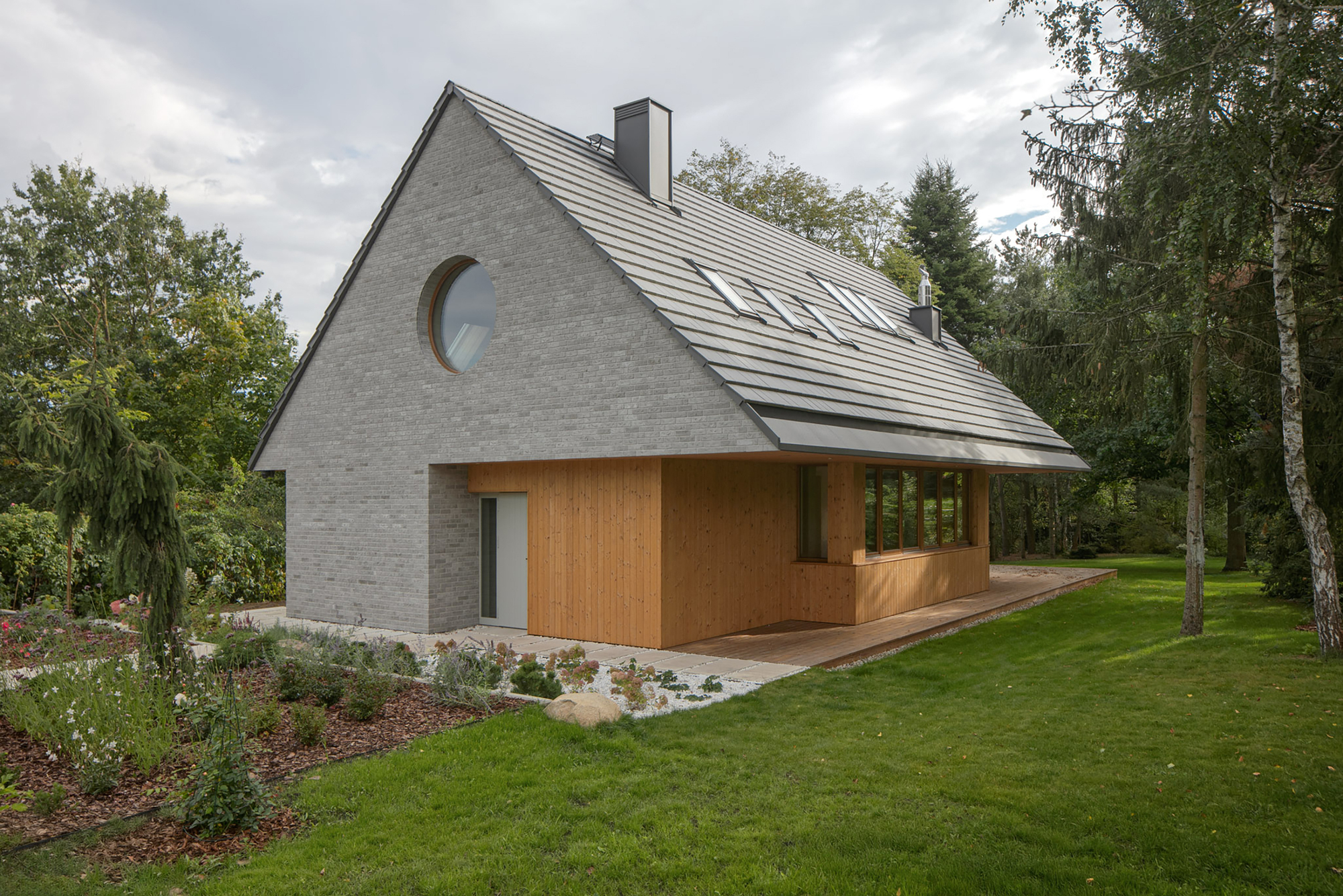 Three-quarter view of the brick gable, round window, timber-clad ground floor and tiled roof with skylights beside a planted garden
