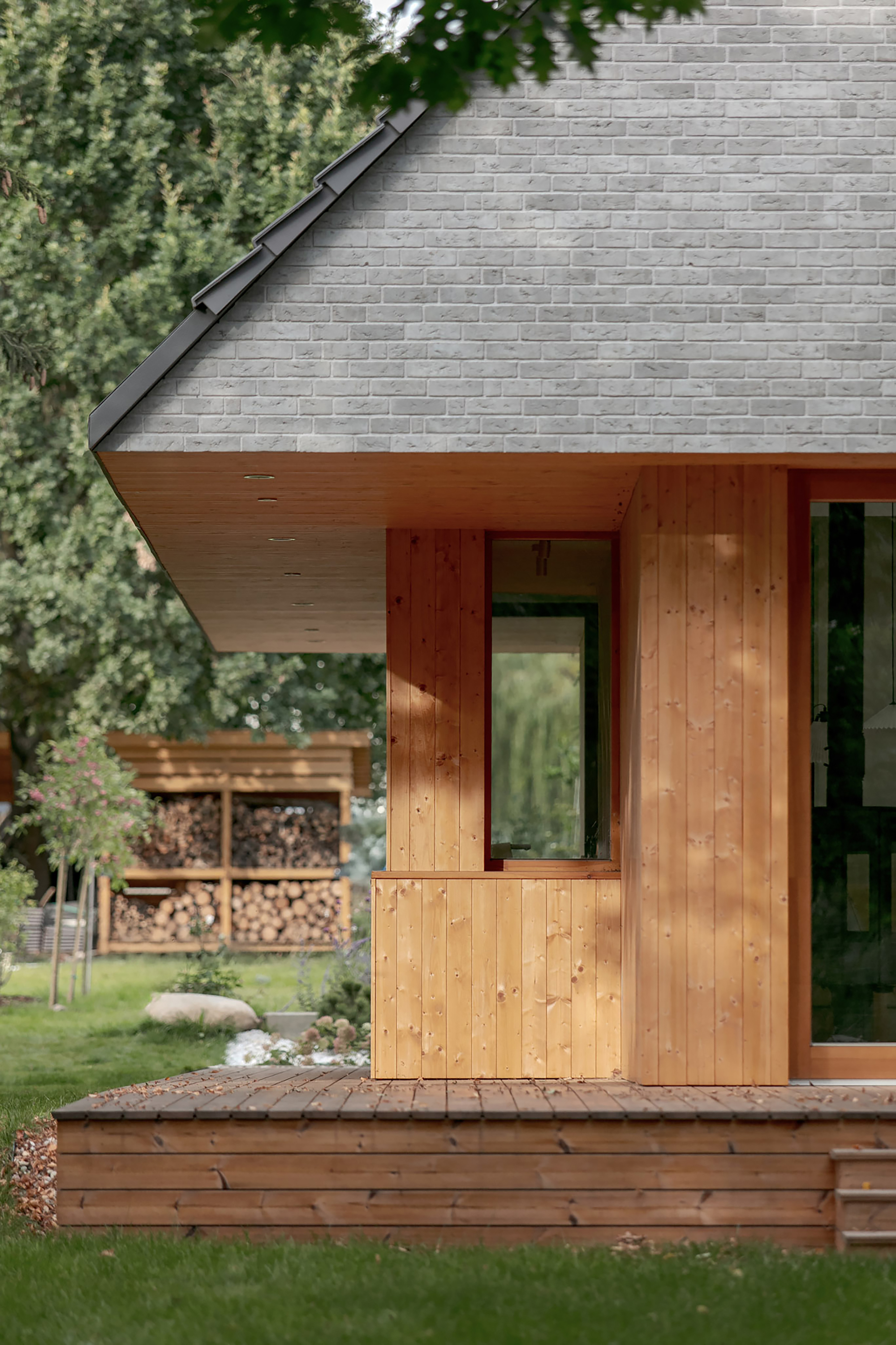 Timber-clad porch corner under the roof overhang, with a stacked firewood shelter glowing in dappled light behind